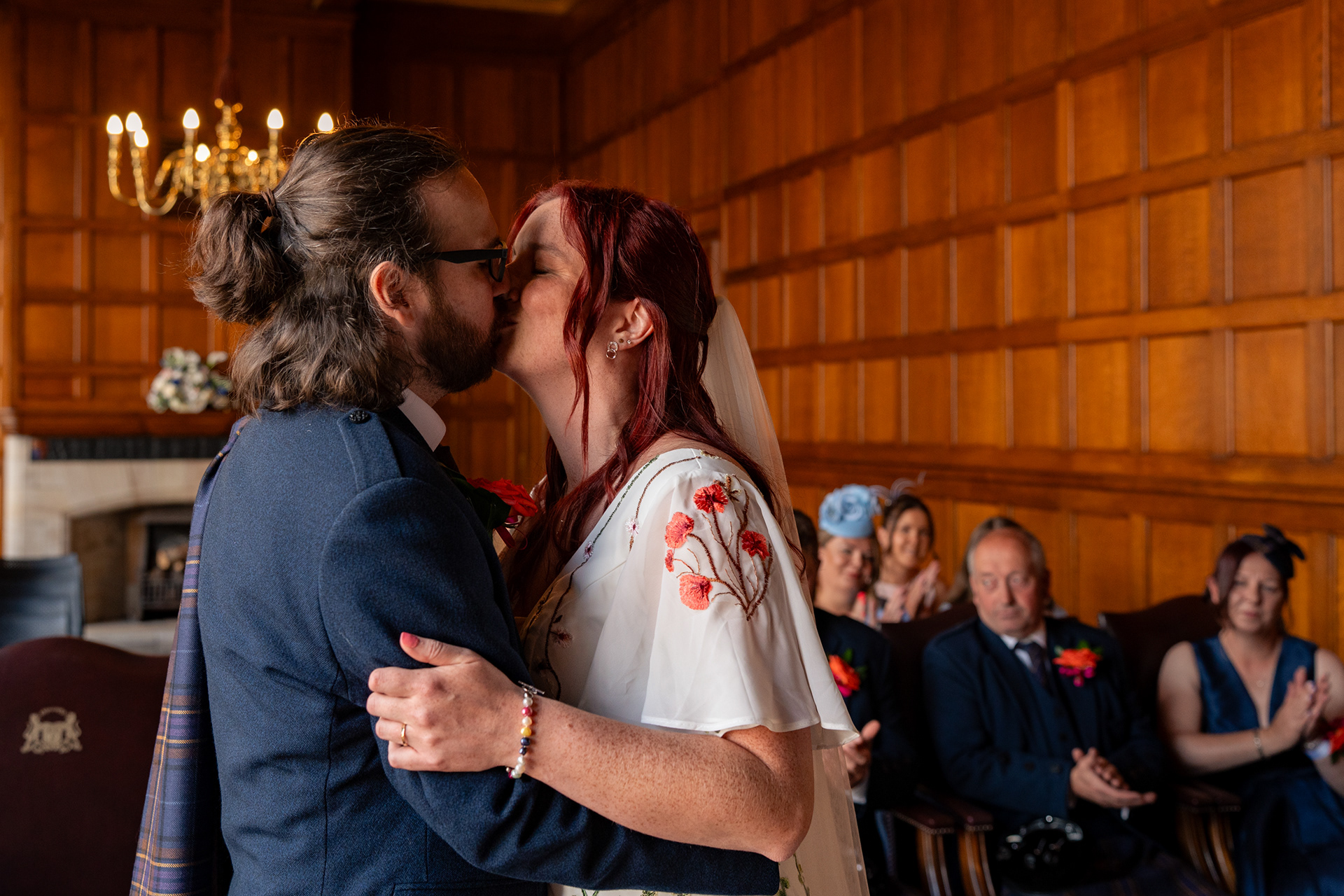 Newlywed bride and groom share their first kiss in the Grant Room at Marischal College. Their guests are visible in the background.