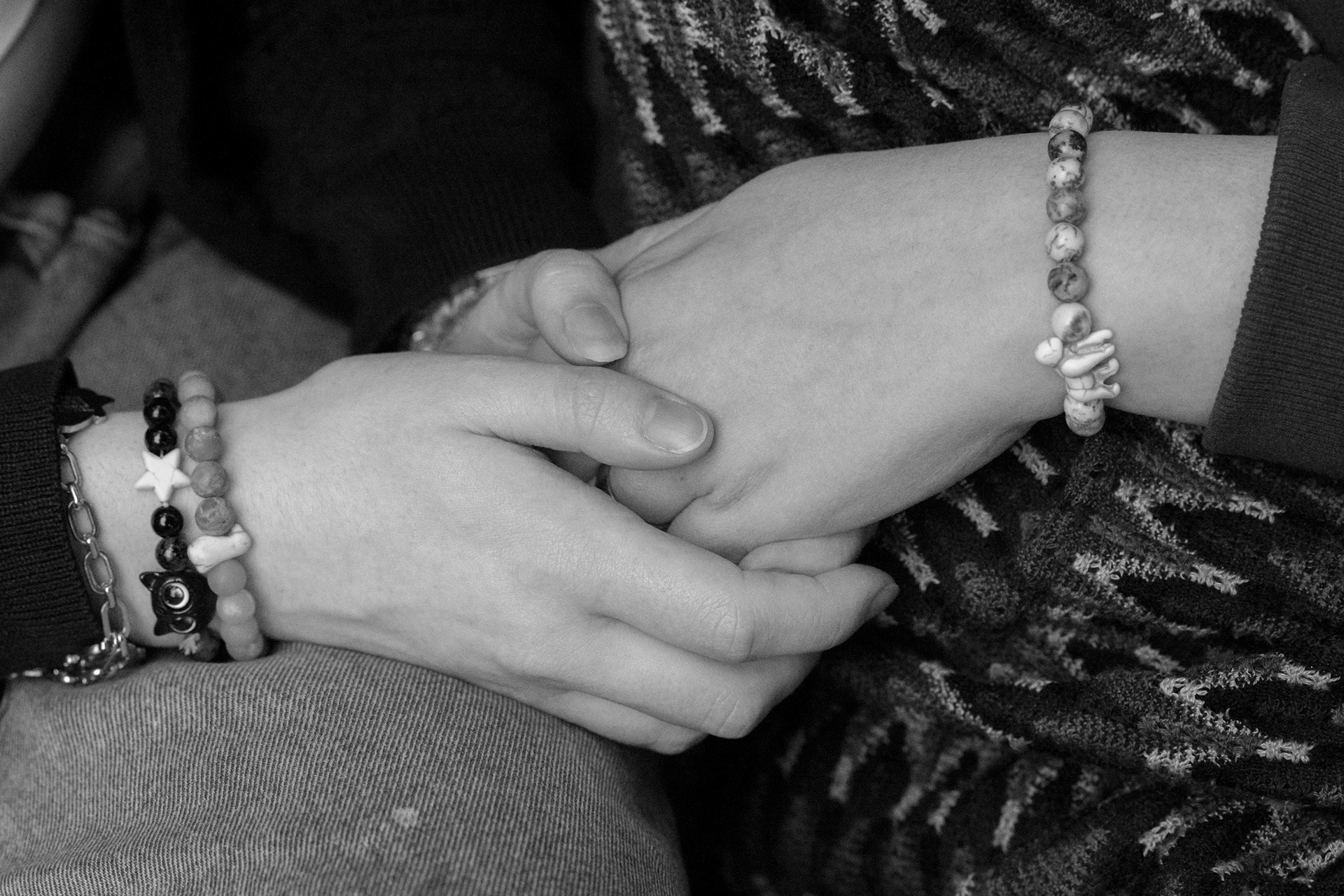 A black and white image of two people's hands clasped together. Both wear beaded bracelets.