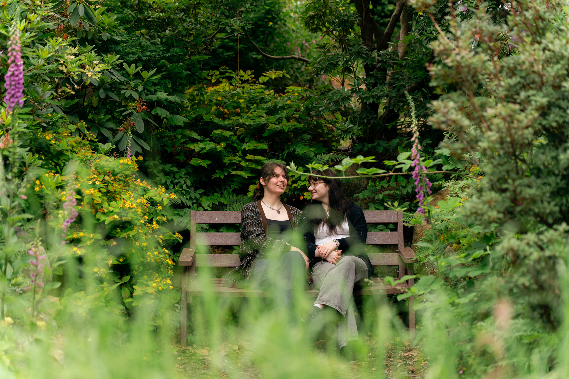 Aberdeen wedding photographer, Sarah, took this photo of a married couple (she/her and they/them) sitting together on a bench, apparently deep in conversation, surrounded and framed by hypericum and foxgloves.