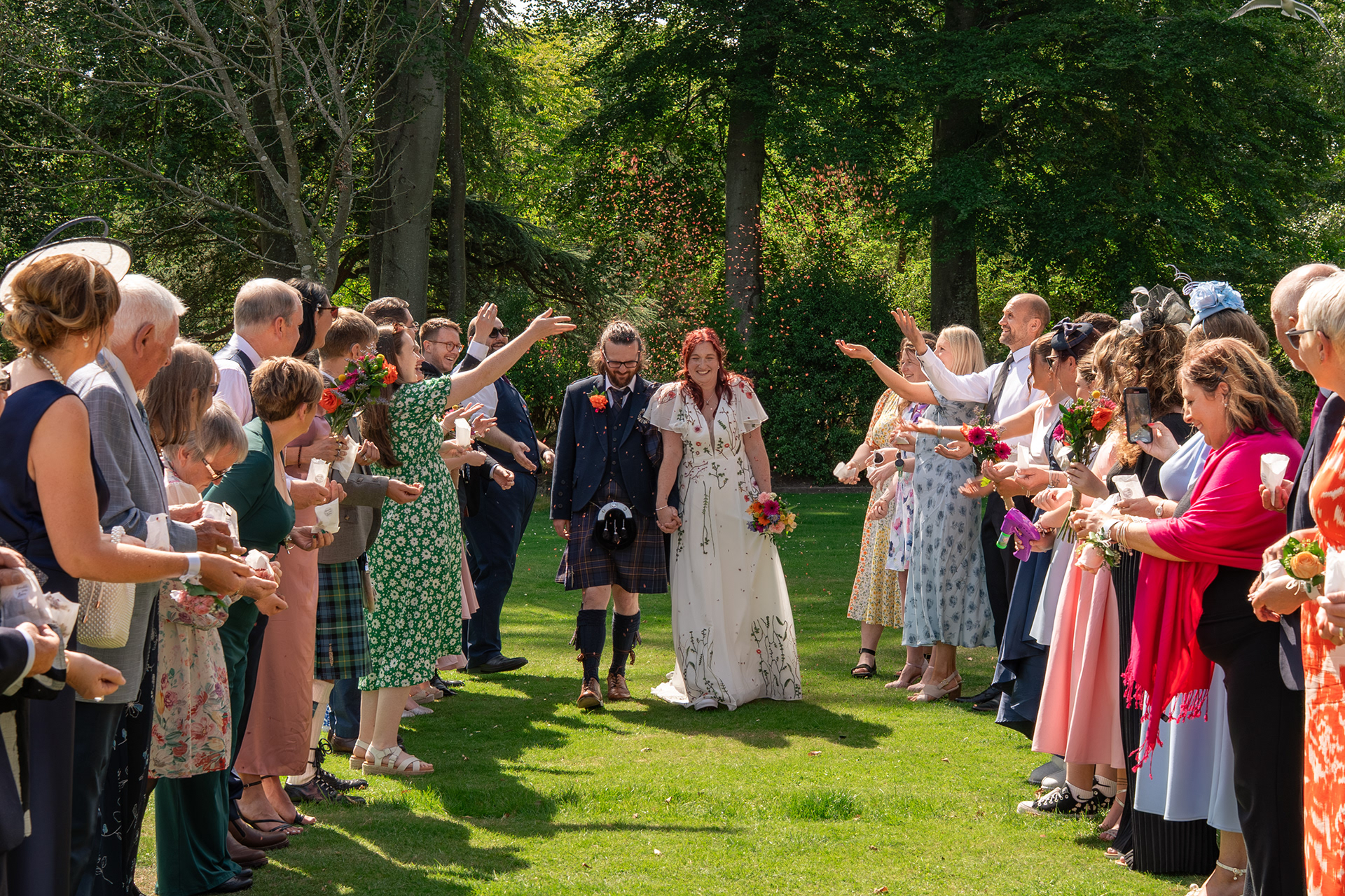 Bride and groom laugh as they are showered with red confetti, walking between their guests on a sunny day in Hazlehead Park