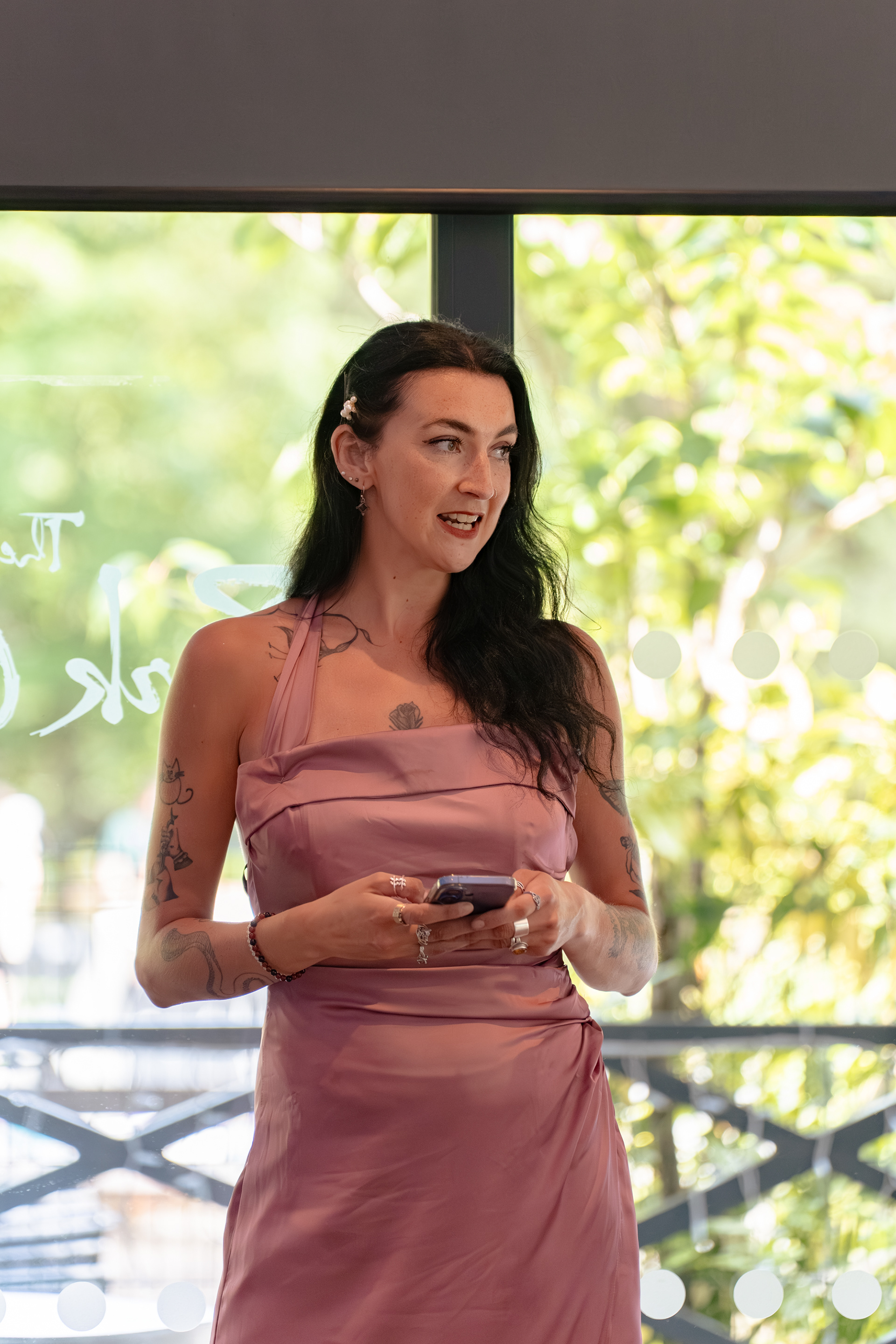 A young tattooed woman with long, dark hair and a dusky pink bridesmaid dress holds her phone and looks ahead as she gives a wedding speech