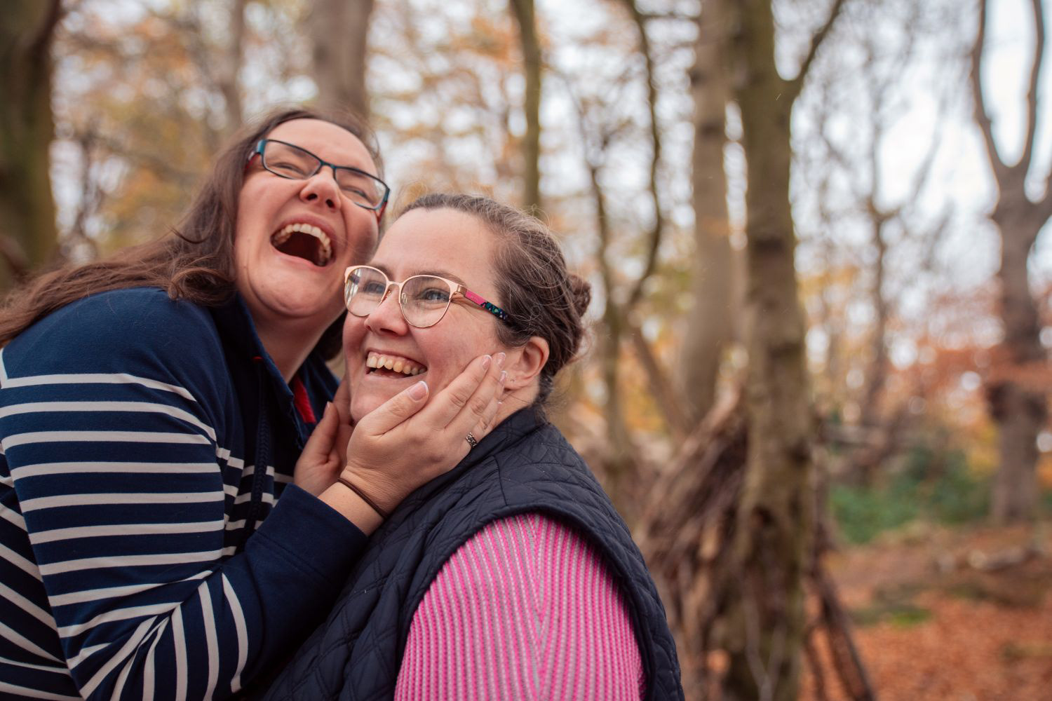 One brunette woman holds her girlfriend's face and screams with loving laughter, in the autumn woods, at their engagement photo shoot.