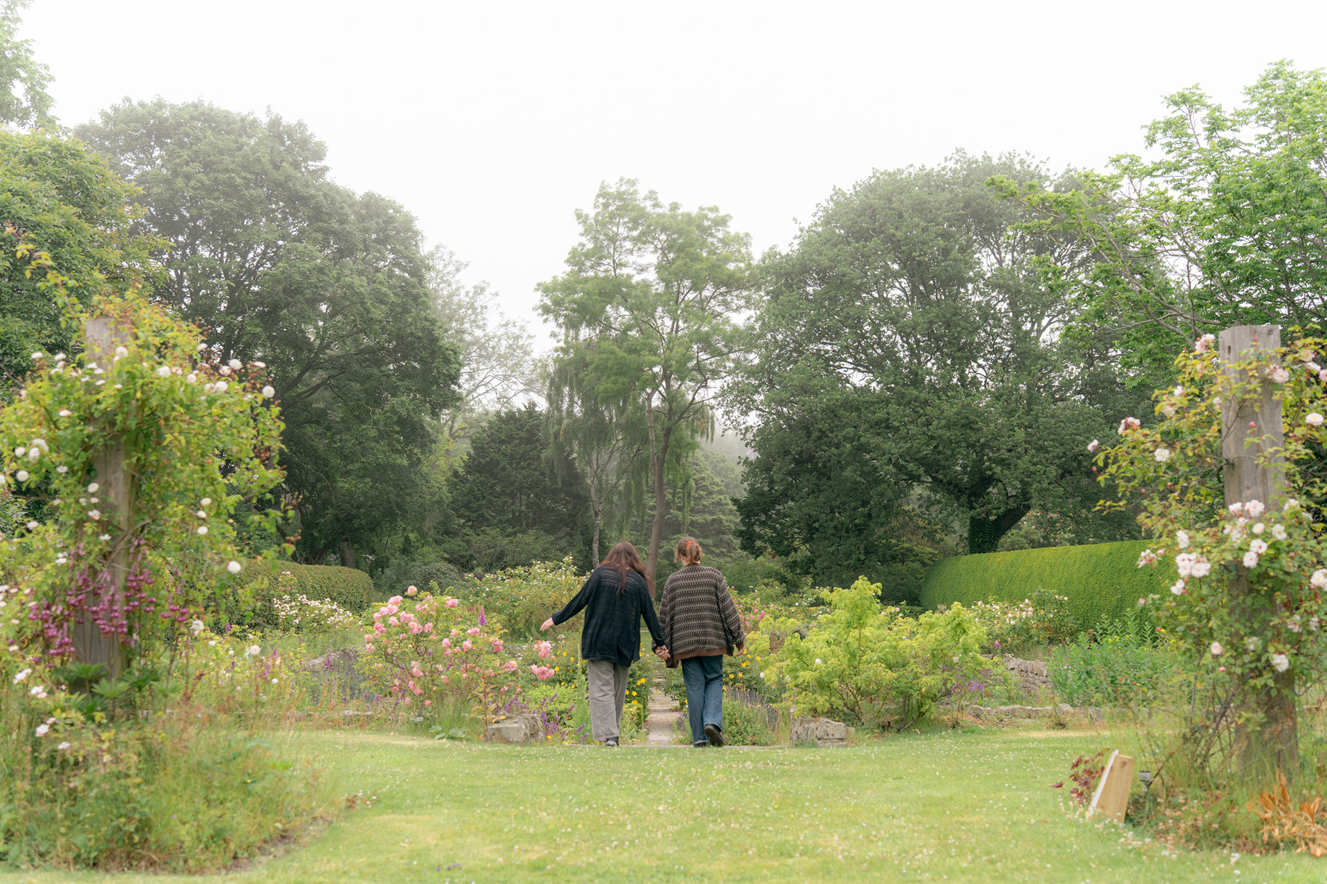 A couple, both in long gardens with long brown hair, hold hands and walk down into a sunken rose garden; the details in the background are soft from the mist which surrounds them.