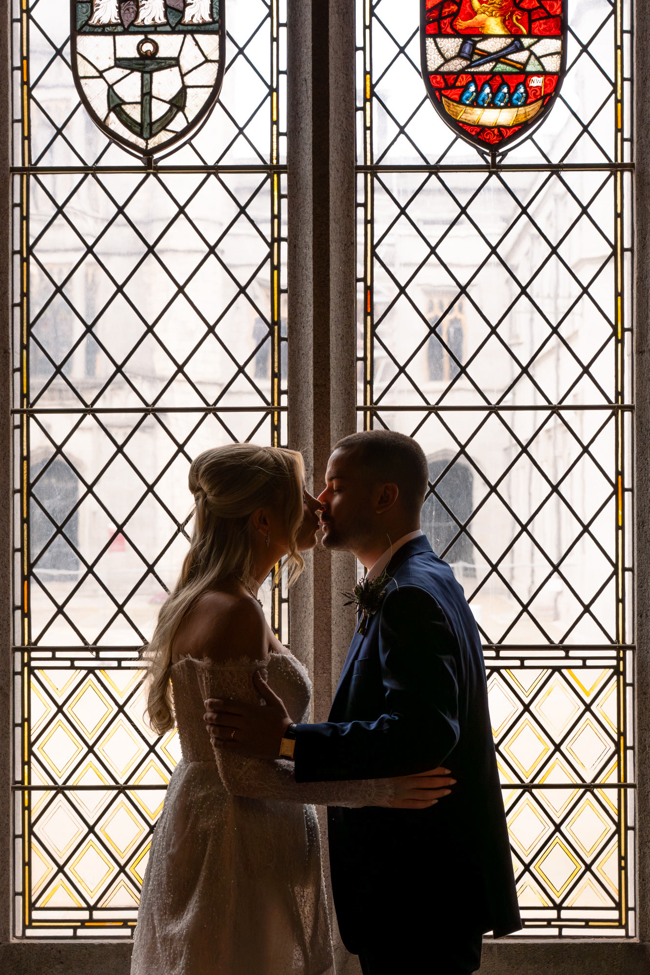 Last Minute Wedding Photographer Aberdeen: A blonde bride in white dress and Spanish groom in blue suit kiss in front of a stained glass window in Marischal College.