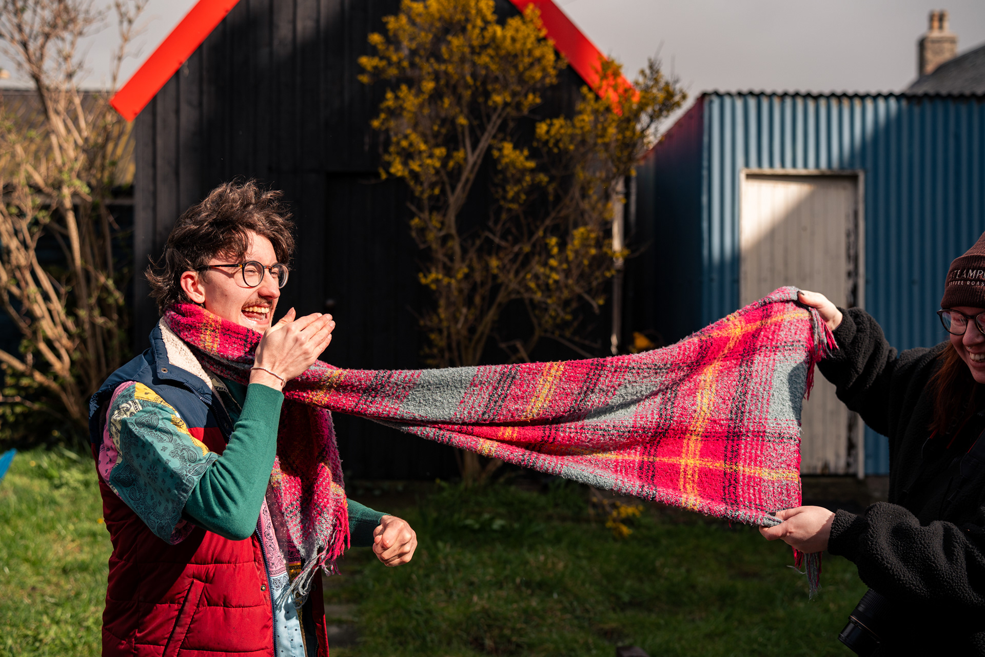 A man in colourful clothes laughs as a woman in black waves his large, bright scarf around beside him.