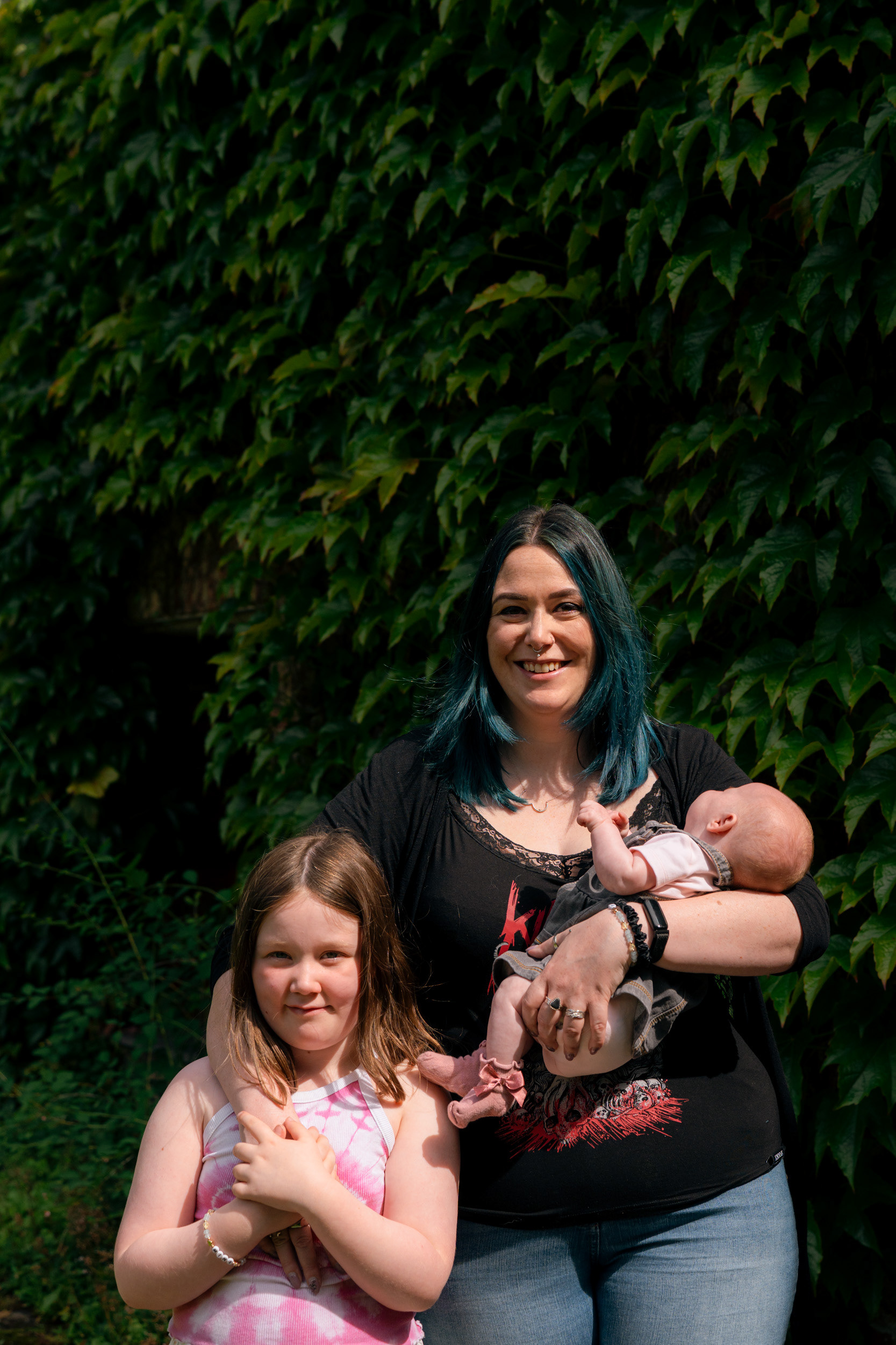 An alternative family photo shoot for two heavy metal fans and their kids. Against a backdrop of Virginia creeper, mum with green hair cradles her newborn daughter in one arm, and wraps her other arm around her 7 year old daughter who is reaching up to hold her hand.