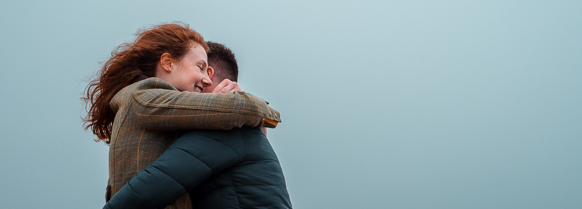 A red-haired woman in a tweed jacket hugs her boyfriend, dressed in a dark blue jacket, against the backdrop of a foggy grey sky. She is beaming. Taken during an Aberdeen couple's photo shoot.