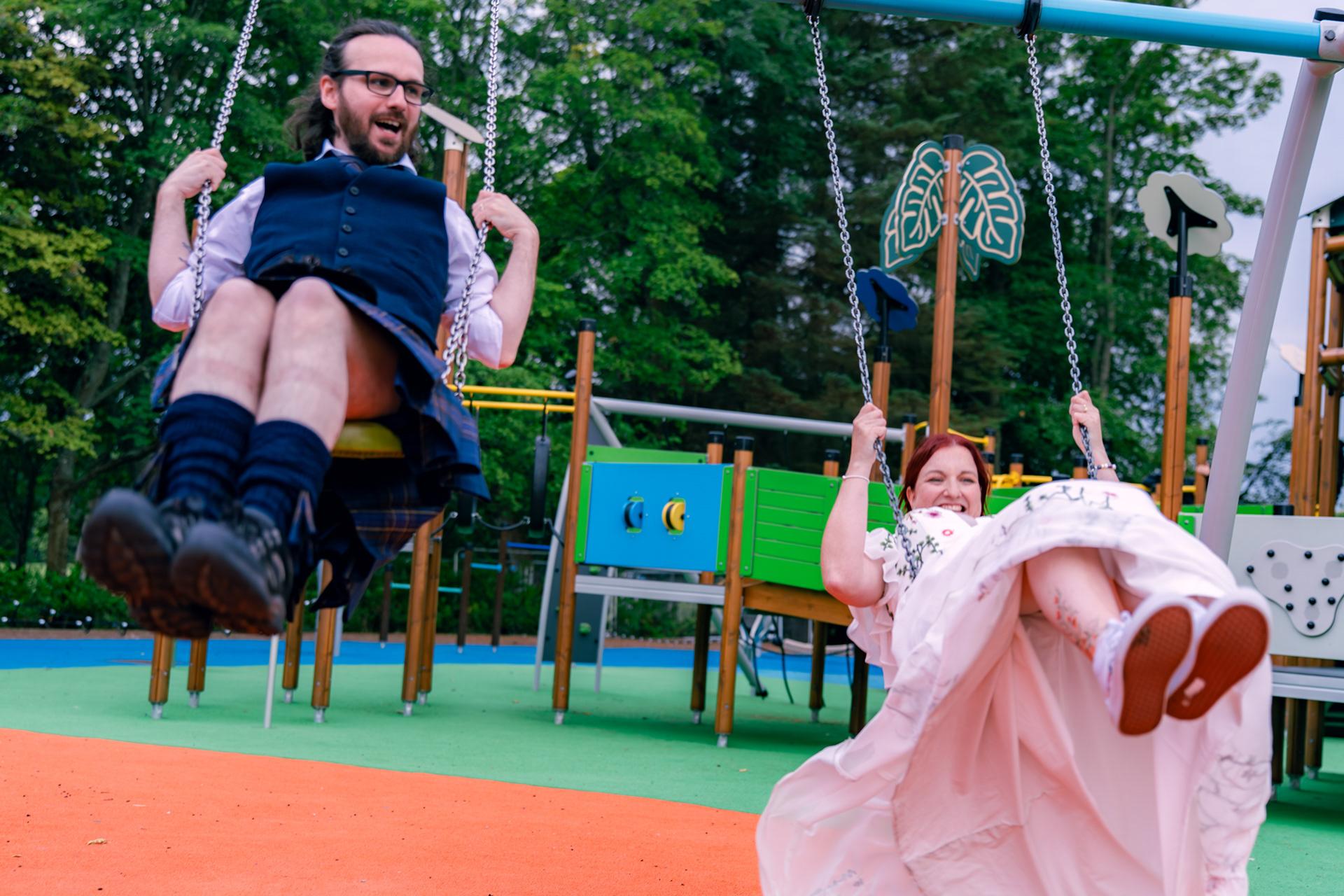 Quirky Aberdeen Wedding Photographer: A groom in blue kilt and bride in floral dress and embroidered Converse laugh as they play on the swings in Hazlehead Park during their wedding reception.