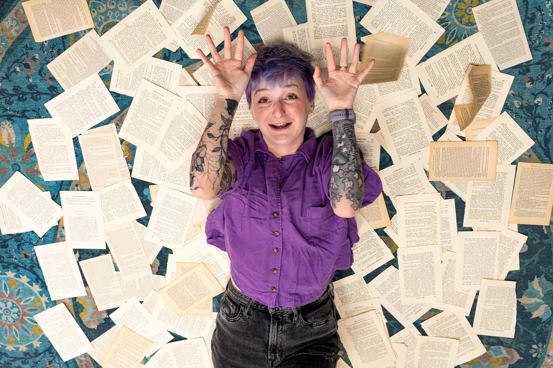 Bookshop owner, Christie, a white woman with short purple hair and amazing bird, cat and floral tattoos laughs as she lies on a floor covered in torn book pages and throws more in the air.