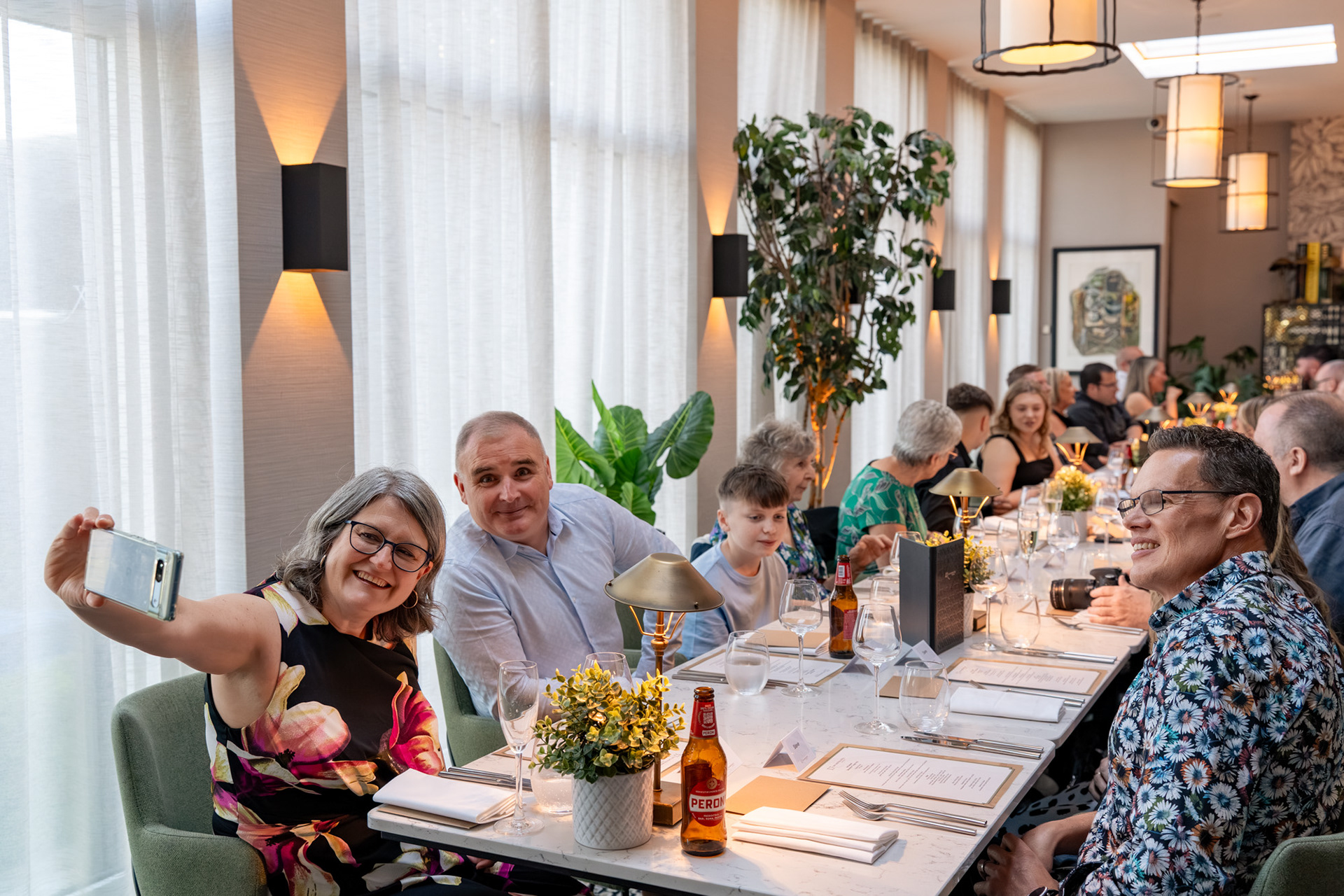 A woman in a black floral dress holds her arm out to take a selfie of people sitting down for a birthday dinner. The photograph shows the length of the table, with around thirty guests chatting along the length of it.