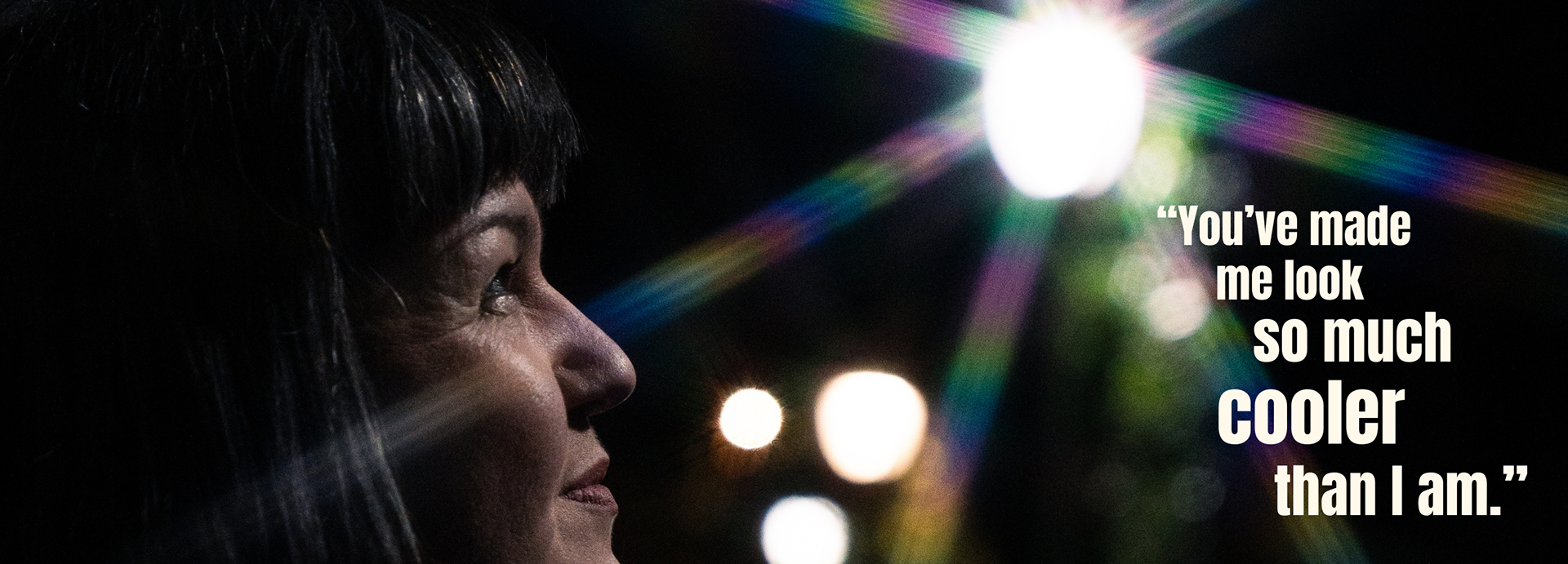 A close up of a woman's face from fringe to lips, side on. In the background, the beams of light from a lamppost are rainbow coloured. The overlaid text reads: "Young woman in pink with tattoos, piercings and short hair blowing in the wind. Review text reads: "You've made me look so much cooler than I am."