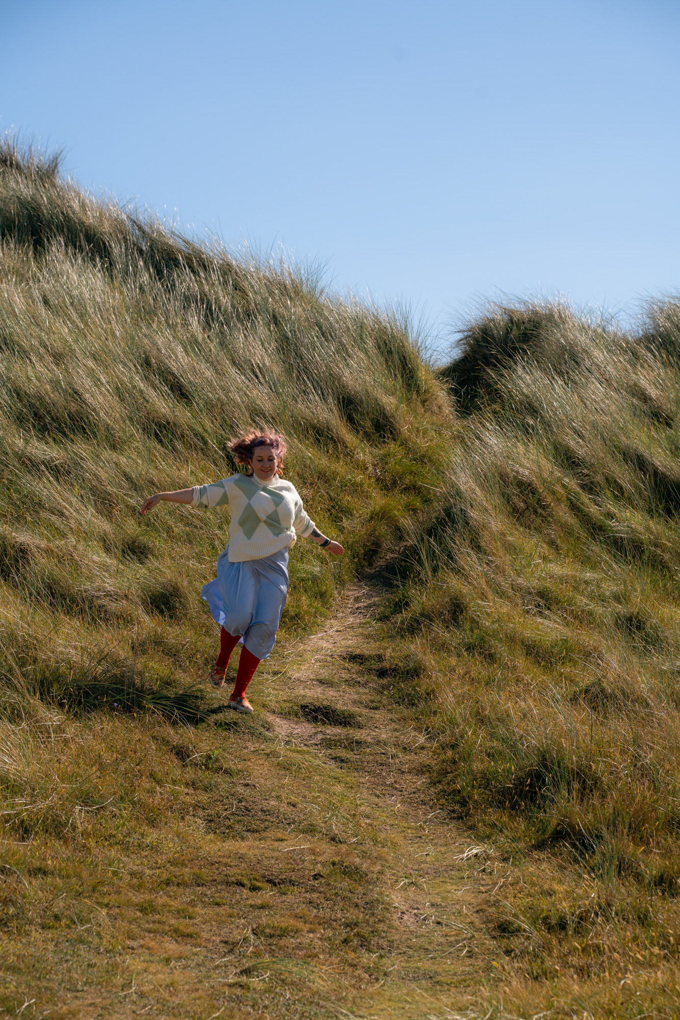 A woman in a white and green jumper and a lilac skirt runs down a grassy slope at an Aberdeenshire beach.