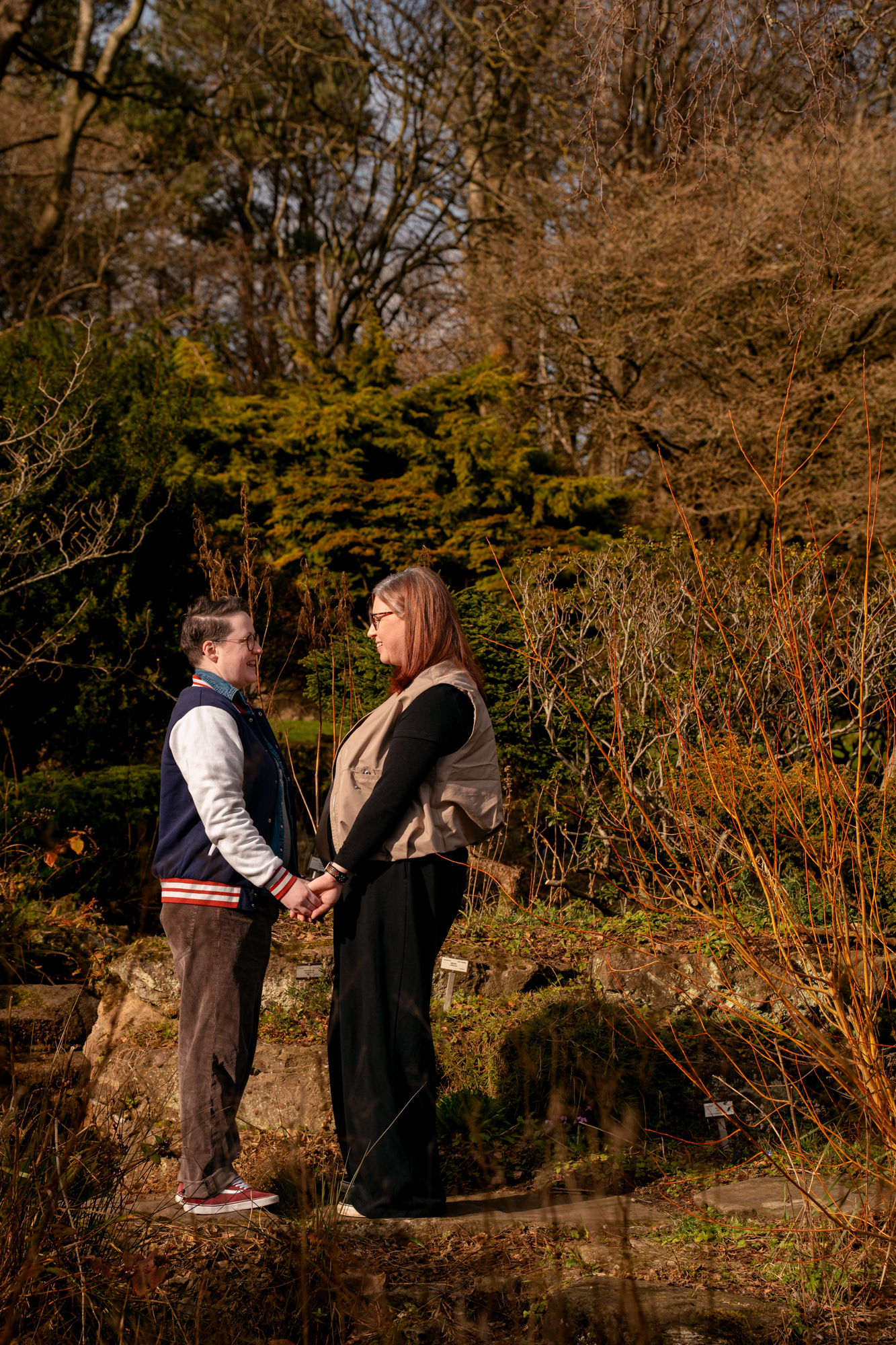 A queer couple (she/her and they/them) stand holding hands, face to face, surrounded by greenery during an Aberdeen springtime photo shoot
