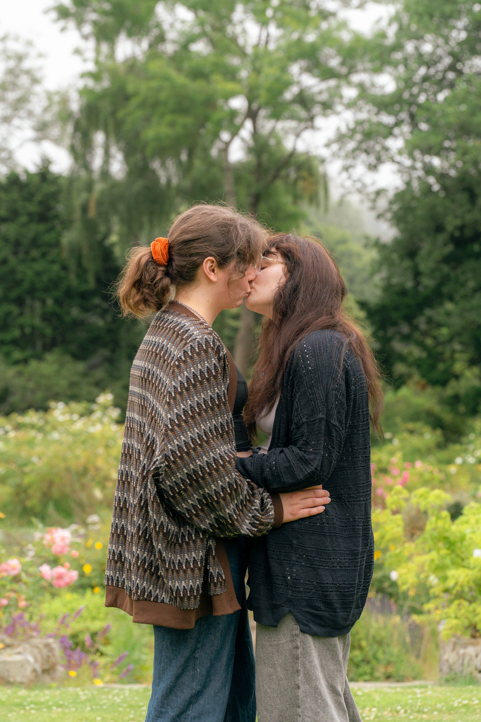 Two wives with long brown hair and long cardigans come in for a kiss, against a backdrop of a misty sunken rose garden in Aberdeen.