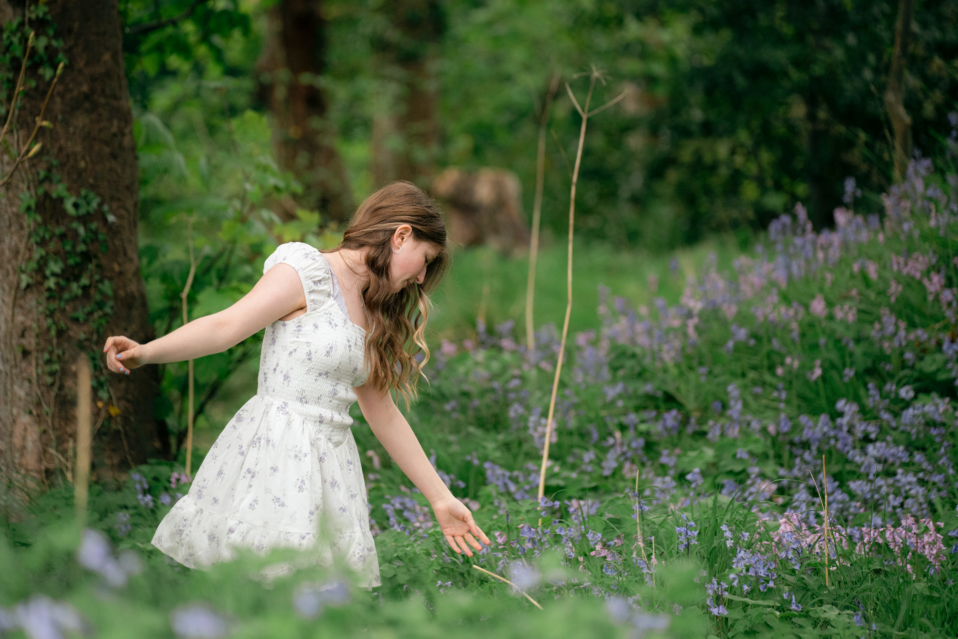 At her Aberdeen family photo shoot, a young woman with long, wavy brown hair, wearing a white floral dress, stands amidst the bluebells with her arms outstretched at she reaches out to touch a flower.