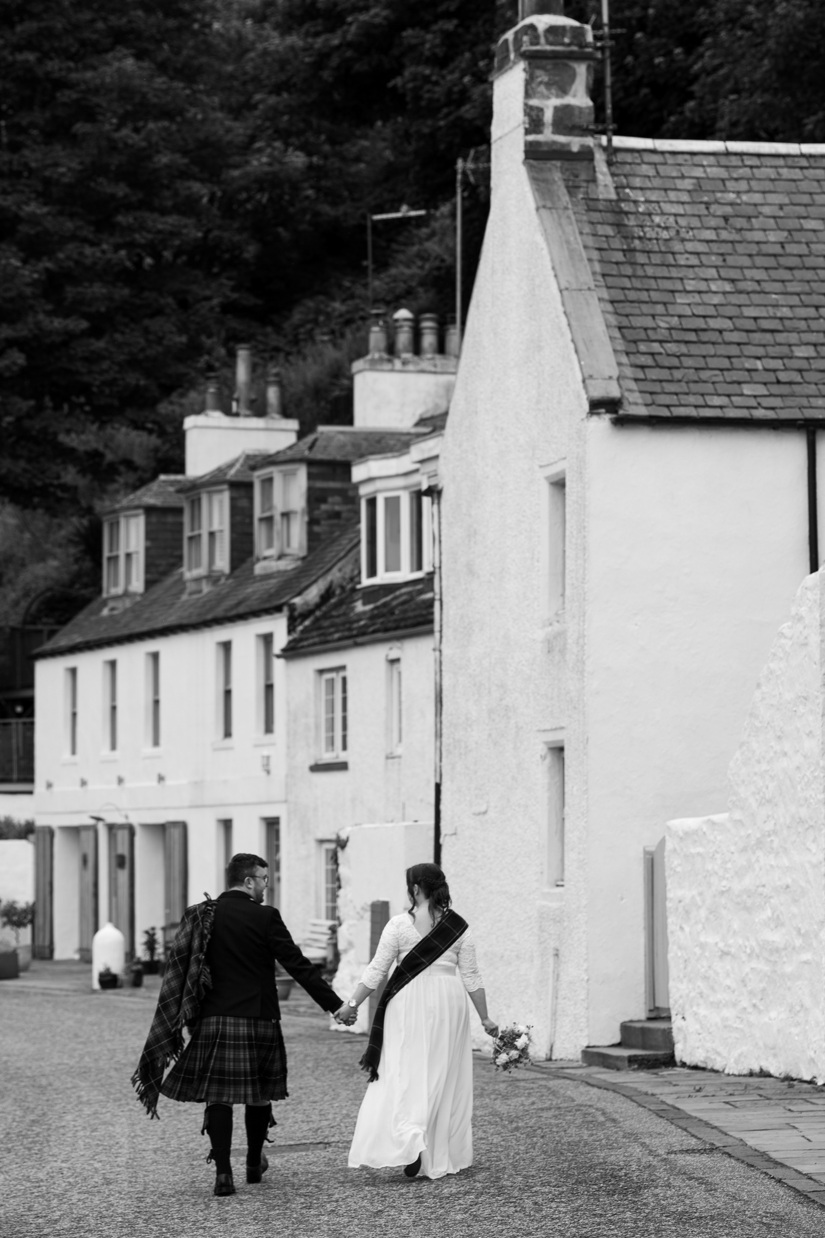 Bride in white dress and groom in kilt walk hand in hand past the wee stone houses at Stonehaven harbour.