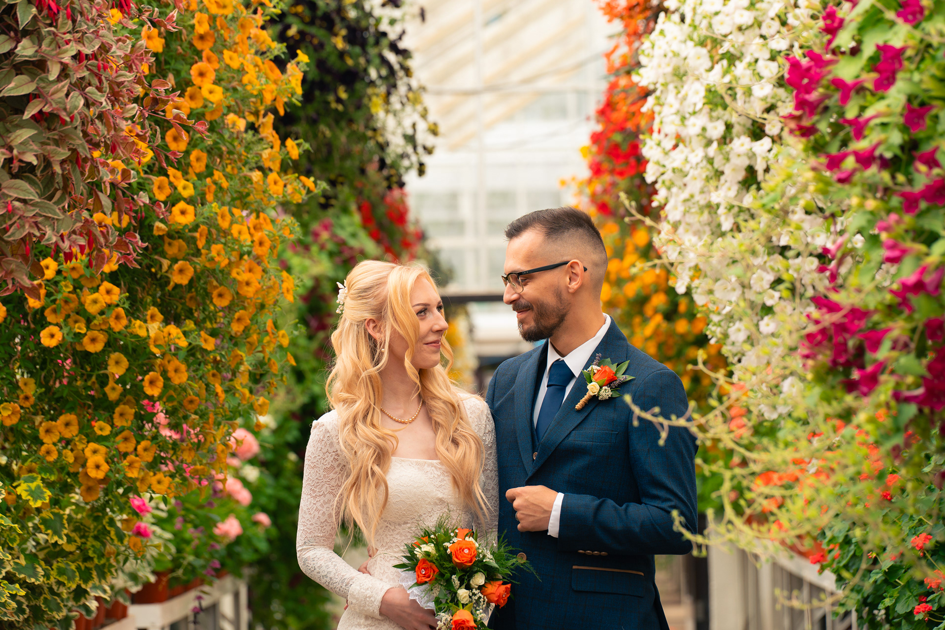 Duthie Park Winter Gardens wedding: Bride and groom smile at each other, surrounded by yellow and orange hanging baskets which complement the bride's orange bouquet.