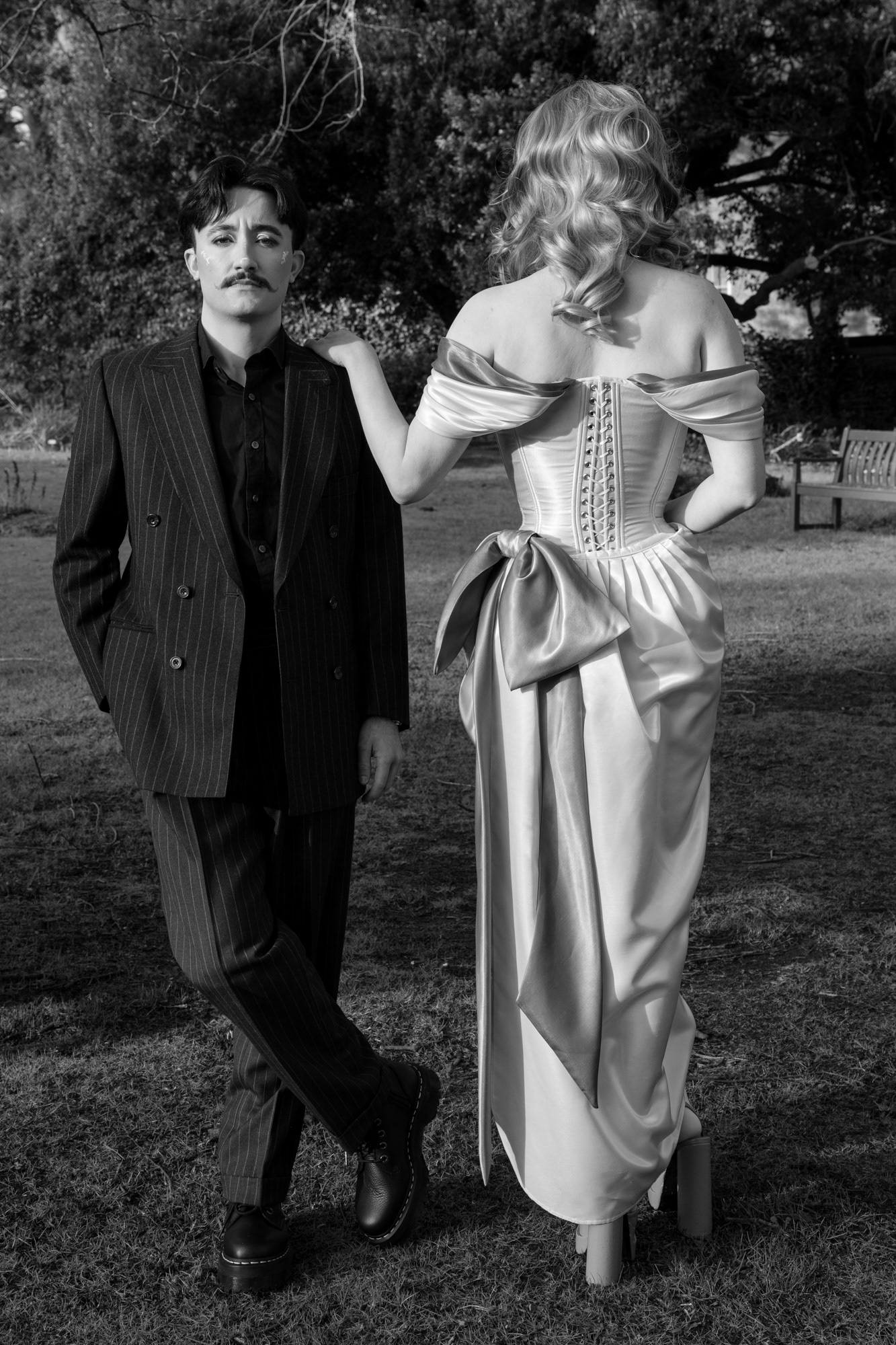 A couple in drag pose together during their Aberdeen couple's photo session. They/them is wearing a pinstripe suit and has short, dark hair and a moustache. She/her is in a glamorous pink and green dress and high green heels with dramatic makeup. They are looking straight at the camera; she has her back turned and one hand on her partner's shoulder so we can see the corset detail at the back of her dress.