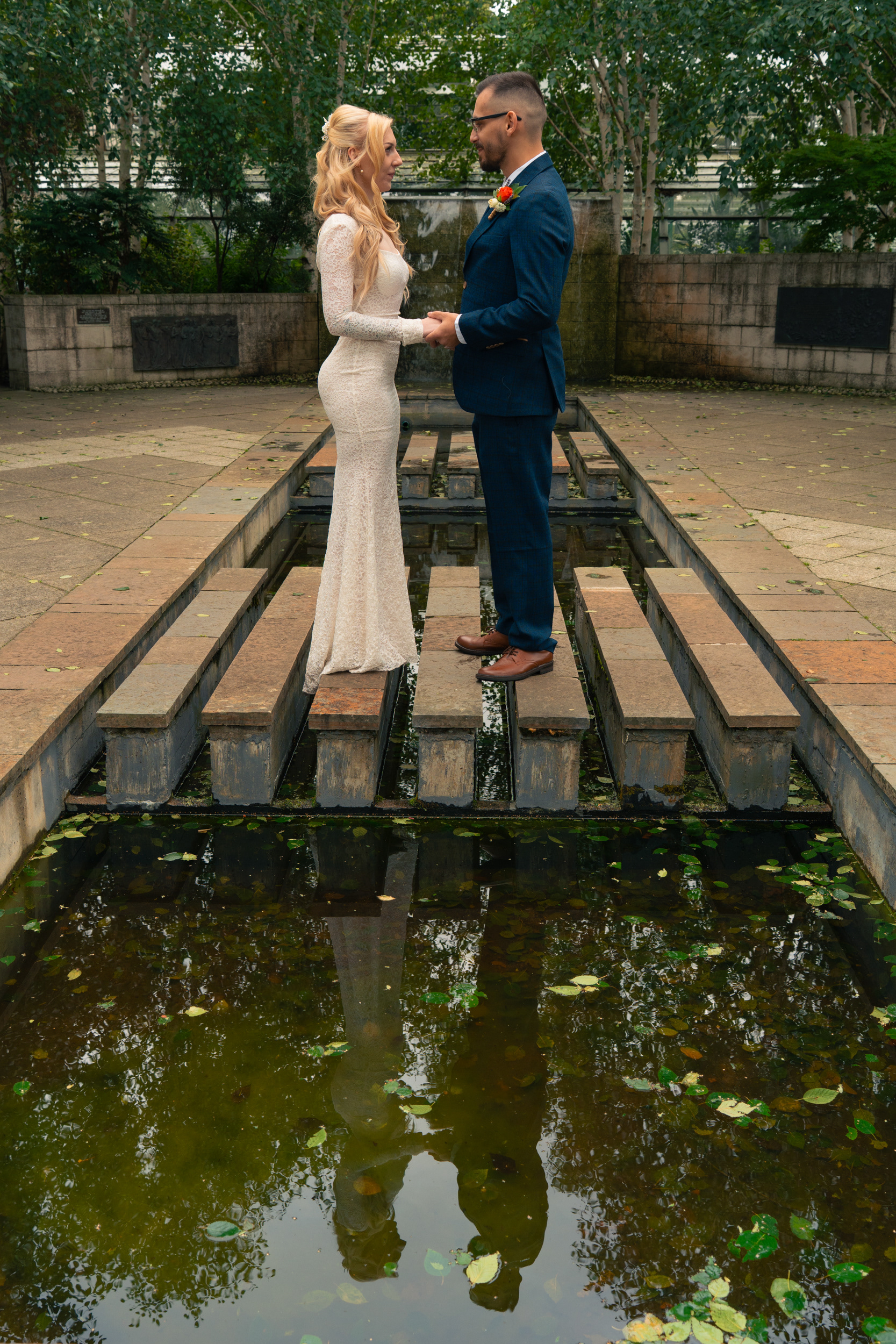 Alternative Aberdeen photography: Bride and groom stand face to face, holding hands, on the stepping stones in Duthie Park. Their reflections are visible in the pool in front of them.