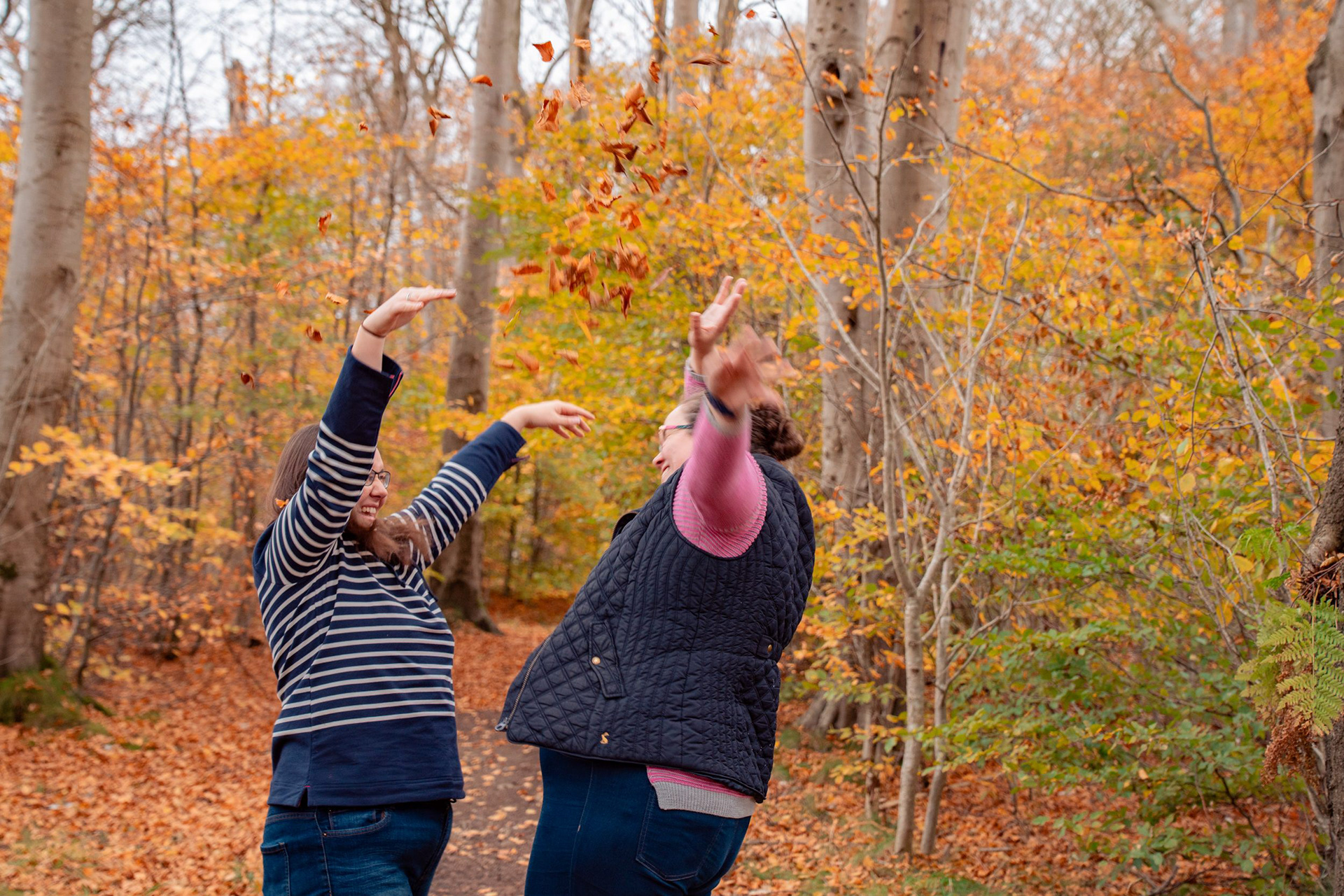Two brunette women throw autumn leaves at each other and laugh during their autumnal Aberdeen pre-wedding photo shoot.