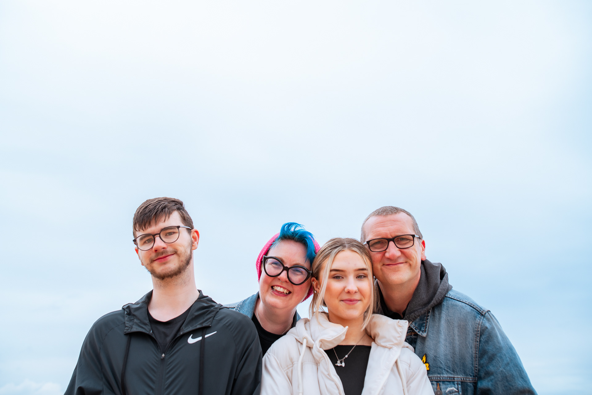 A family of four - two adults, two teenagers - smile directly at the camera with nothing but blue sky behind them