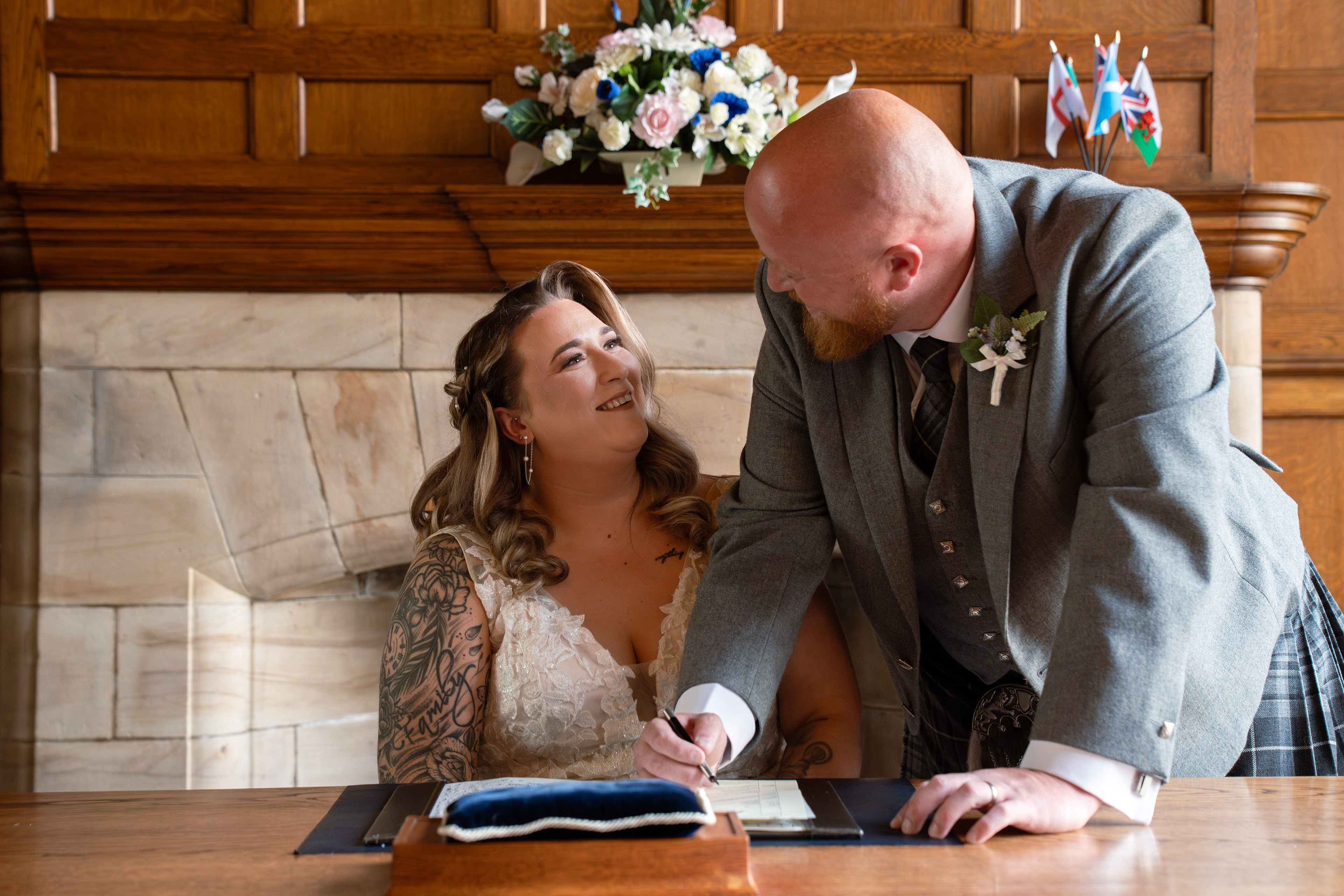 Beautiful tattooed bride in white beams up at her new kilted husband as he signs the register
