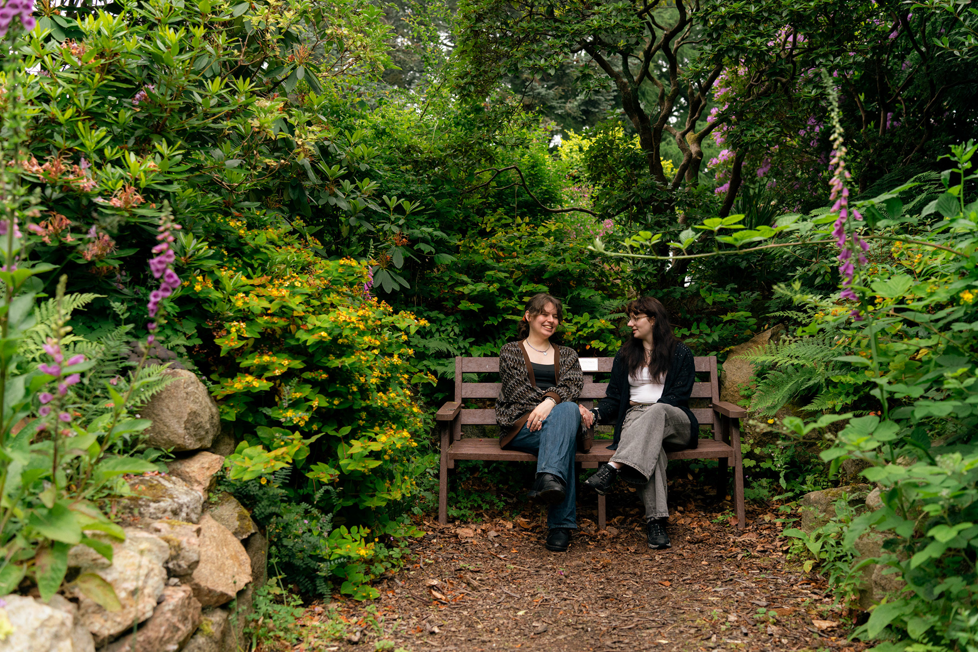 A married couple (she/her and they/them) both in jeans and long cardigans, sit and laugh together on a bench, completely surrounded by hypericum, foxgloves and rhododendrons. Photo taken during their couple's photo shoot with Aberdeen photographer, Sarah.