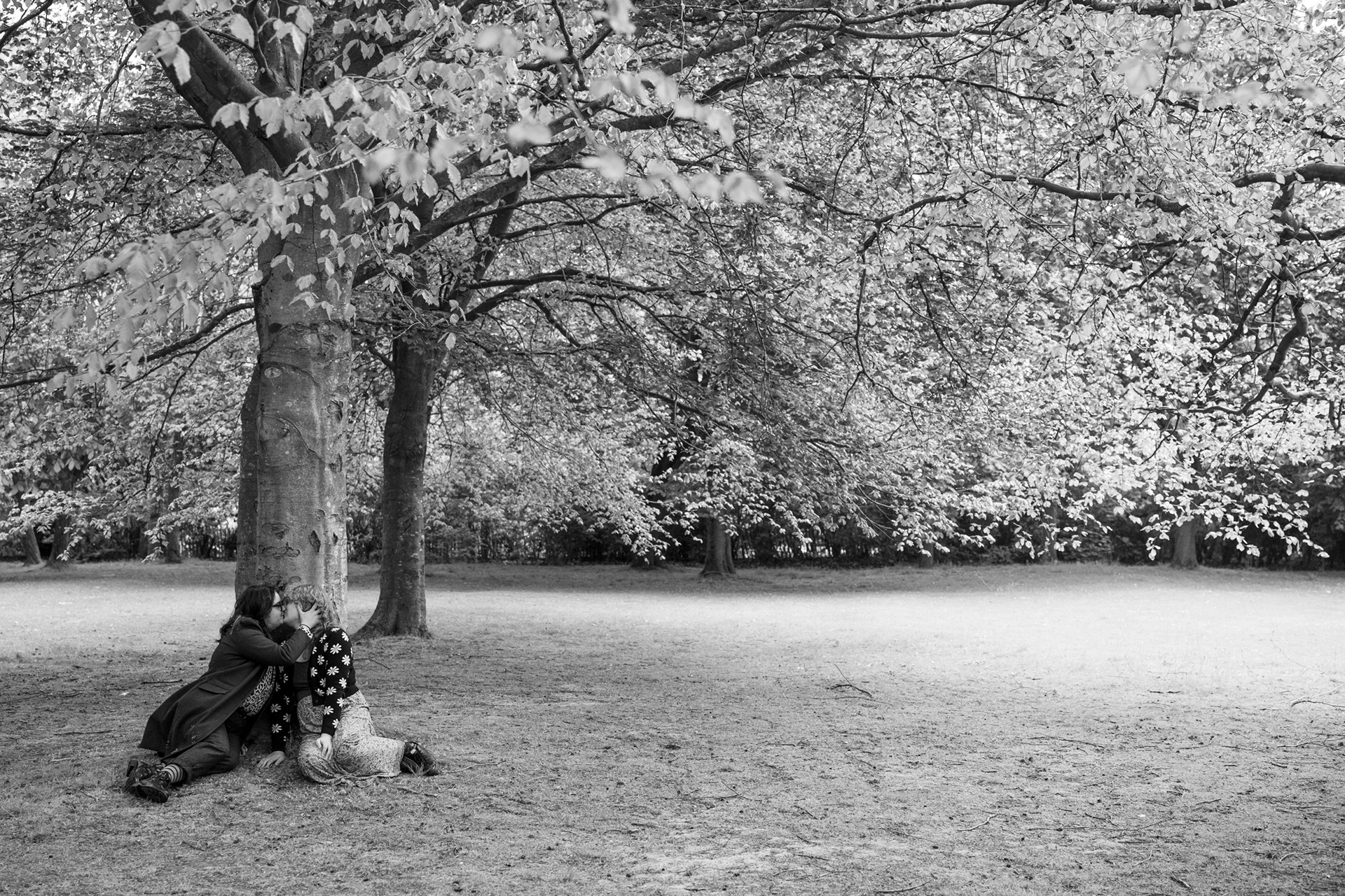 Taken by Scottish LGBTQ+ photographer, Sarah at Windswept Stories, this black and white image shows a queer couple (he in a long coat; she in a floral dress and cardigan) kissing under a sycamore tree with a backdrop of more trees glowing behind them.