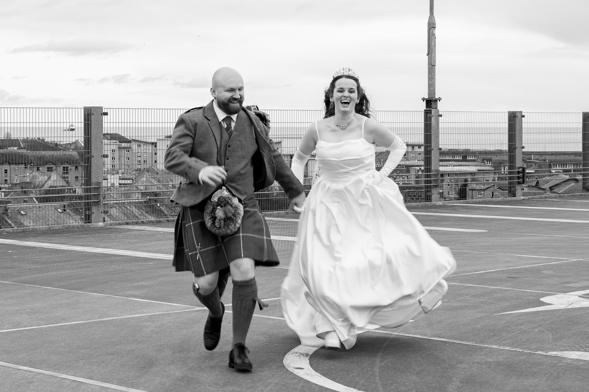 Alternative Aberdeen Wedding Photography: Newlywed couple hold hands and laugh as they run across a car park towards the camera.