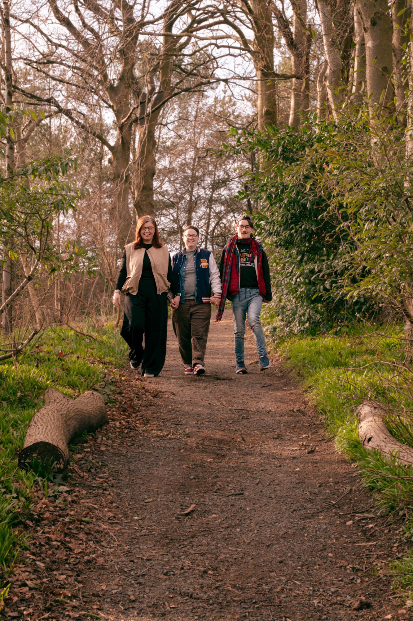 A queer couple (she/her and they/them) and Disabled man (he/him) walk together, all holding hands, down a woodland path.
