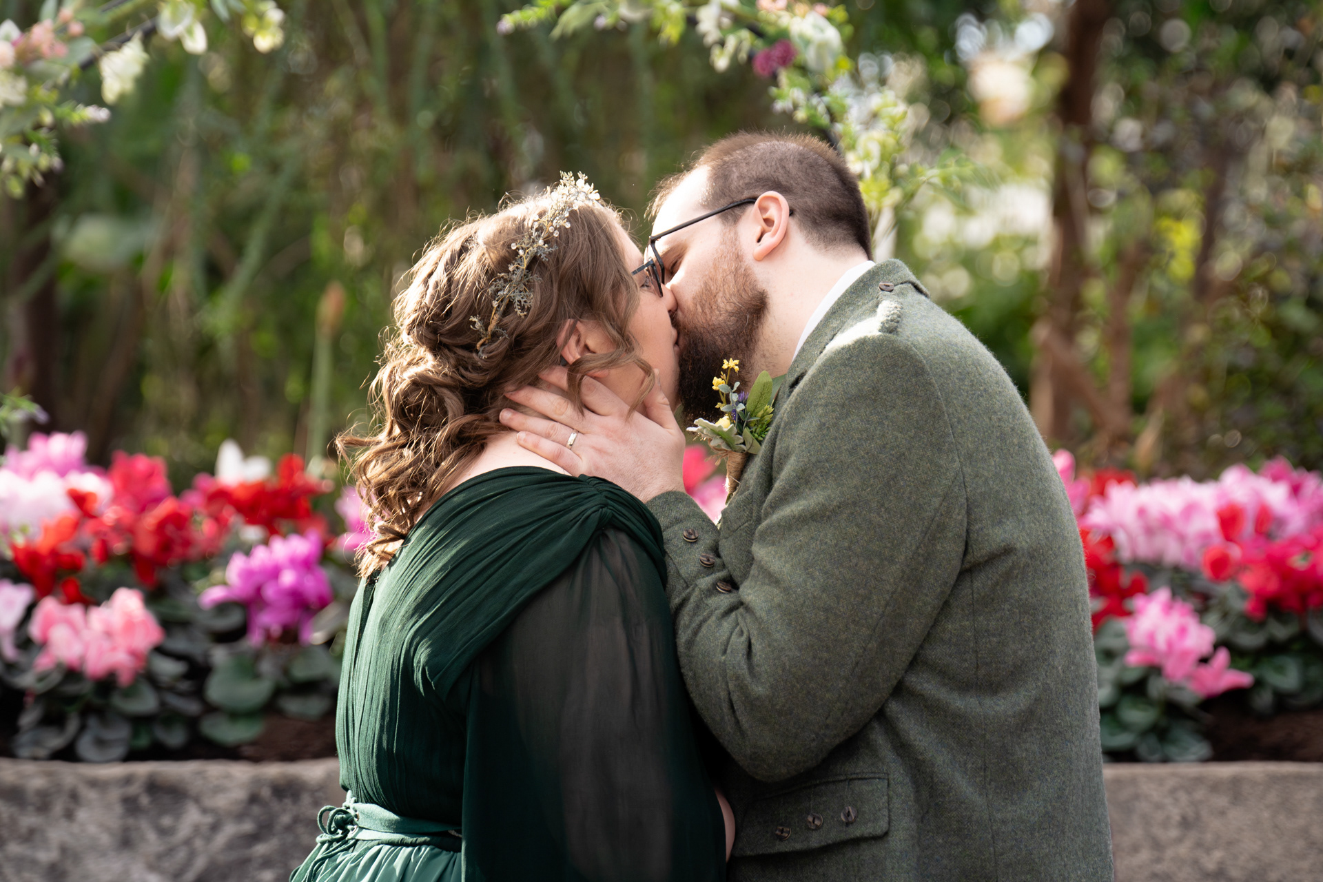 A bride in a green dress with flowers in her hair is kissed passionately by her husband in green tweed, during their Duthie Park wedding. The background is pink and red cyclamen.