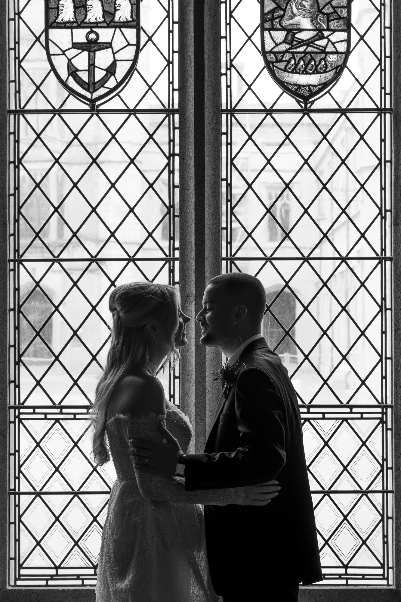 A black and white photo: A newlywed couple lean in for a kiss in front of a stained glass window; they are largely silhouetted with highlights around their faces.
