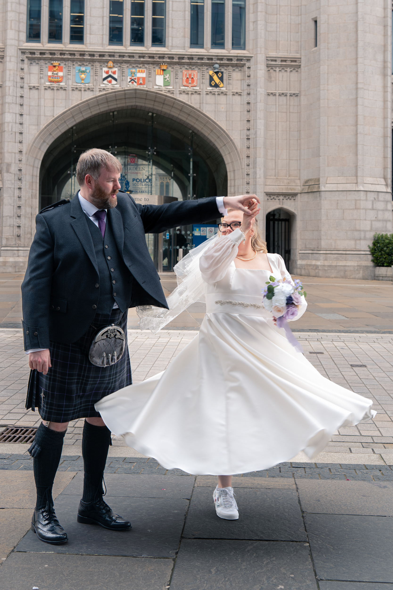 Groom in kilt spins bride in white dress and trainers outside Marischal College
