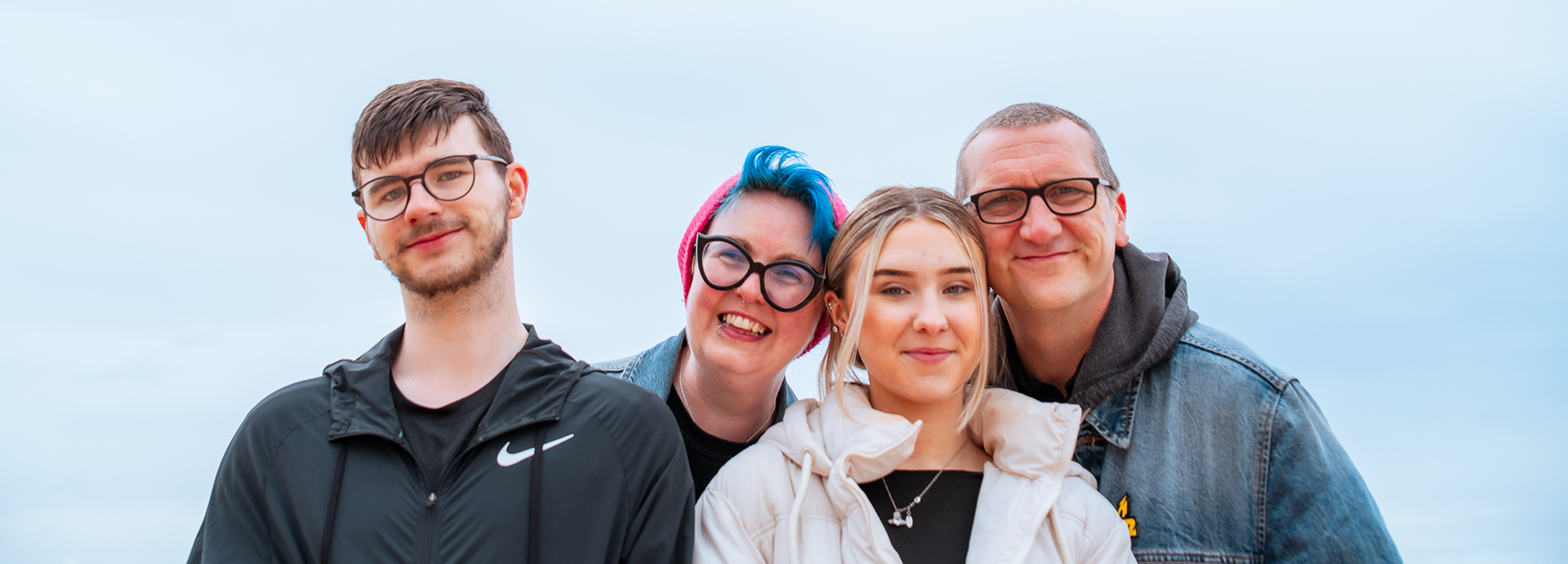 A family of four - two adults (one a woman with blue hair) and two teenagers - smile straight into the camera lens at their Aberdeen family photo shoot. The background is entirely blue sky.