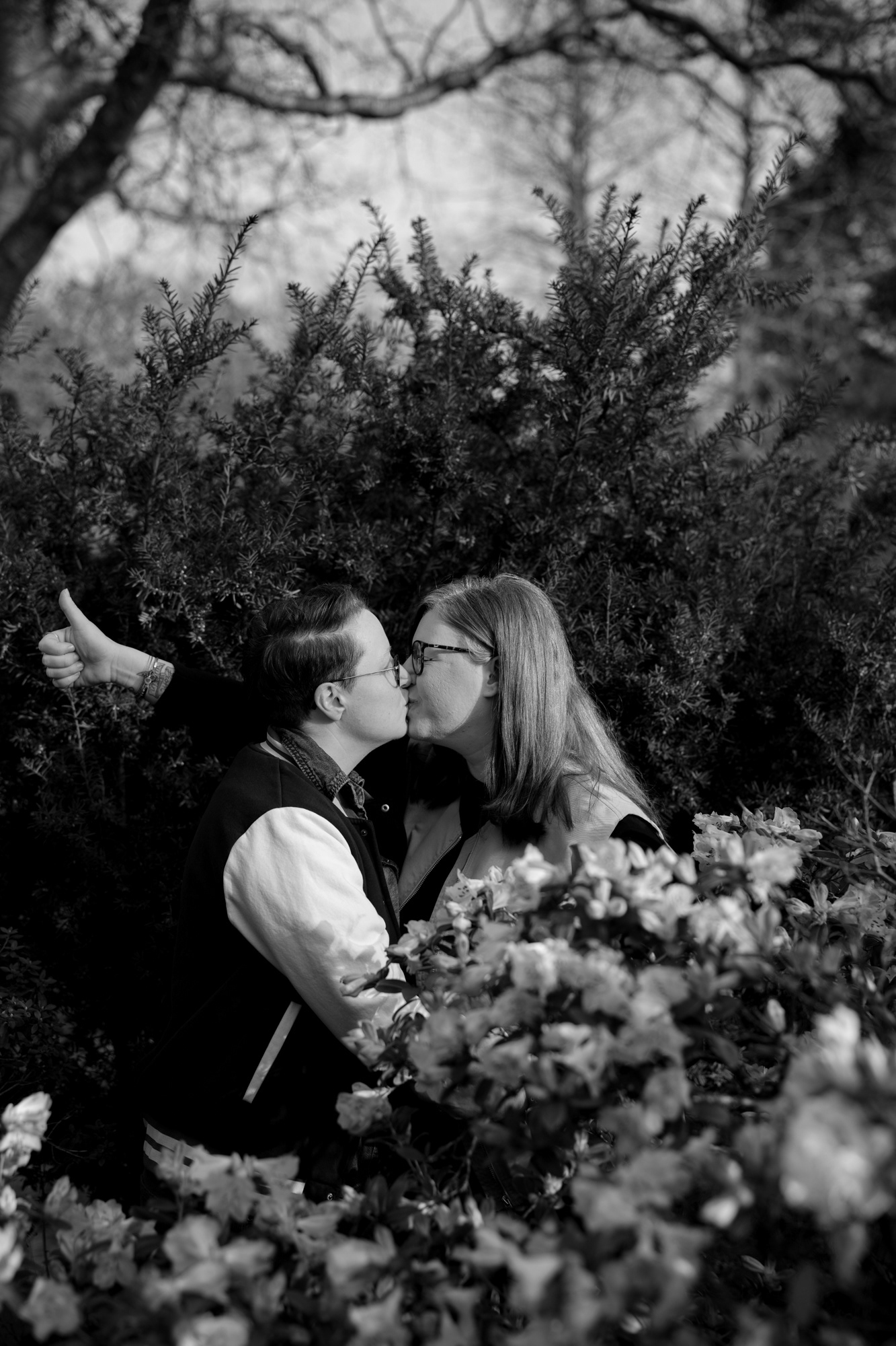 A queer couple (she/her and they/them) kiss amongst the spring blossom, one doing a thumbs up behind her partner's back