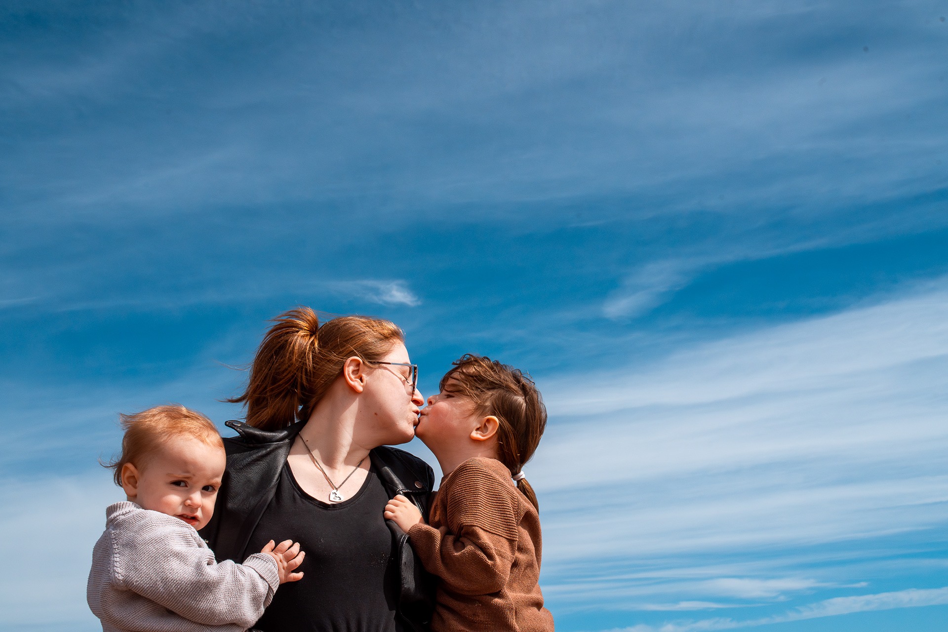 A red haired mum in a leather jacket holds her baby, who is looking at the camera, and young son, who she is kissing. The background is entirely blue sky. A gorgeous day for this family photo shoot on Aberdeen beach.