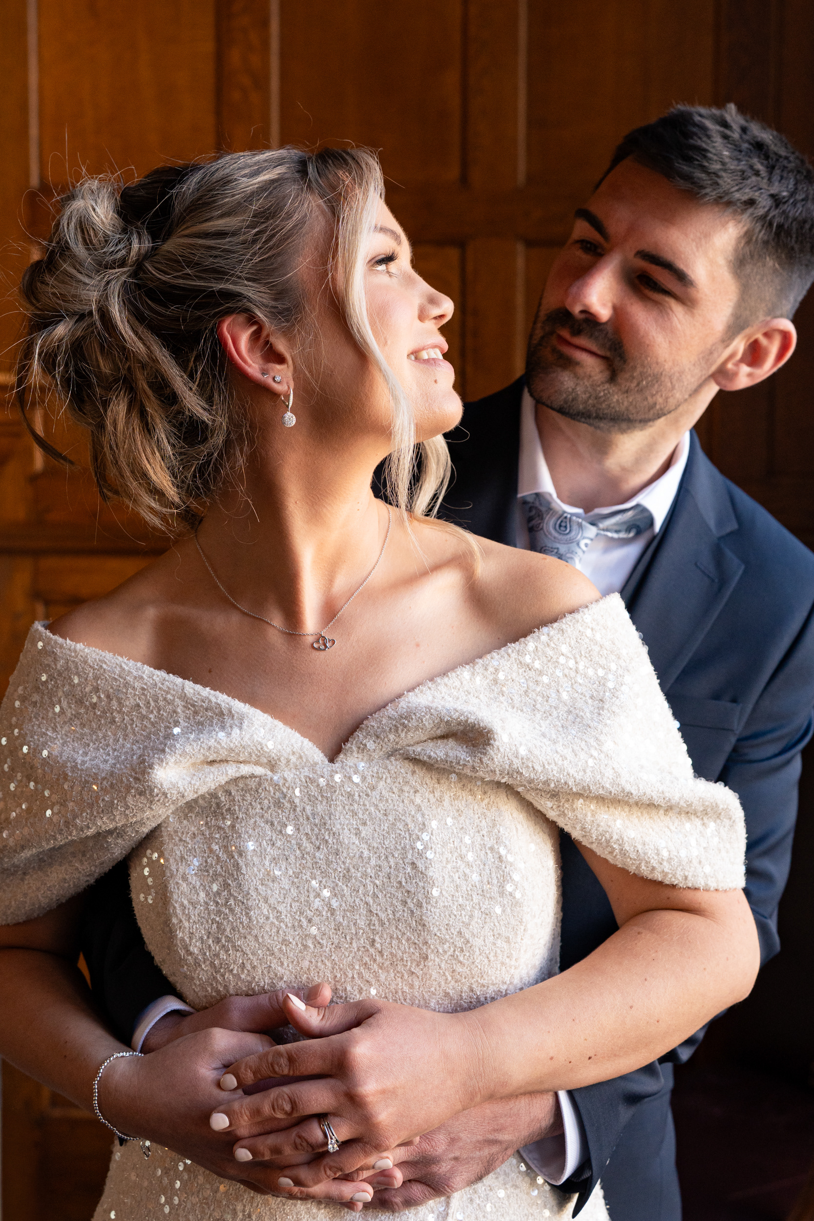 A blonde bride in sparkly dress turns to smile at her new husband in blue suit, side lit by a window.