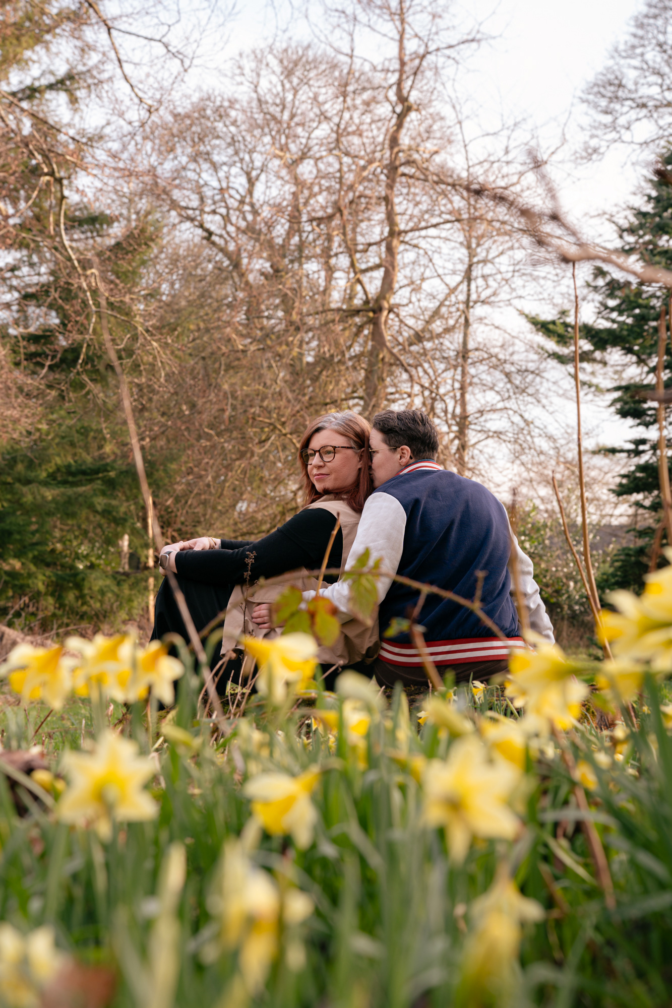 A queer couple (she/her and they/them) sit and kiss behind a wall of daffodils.