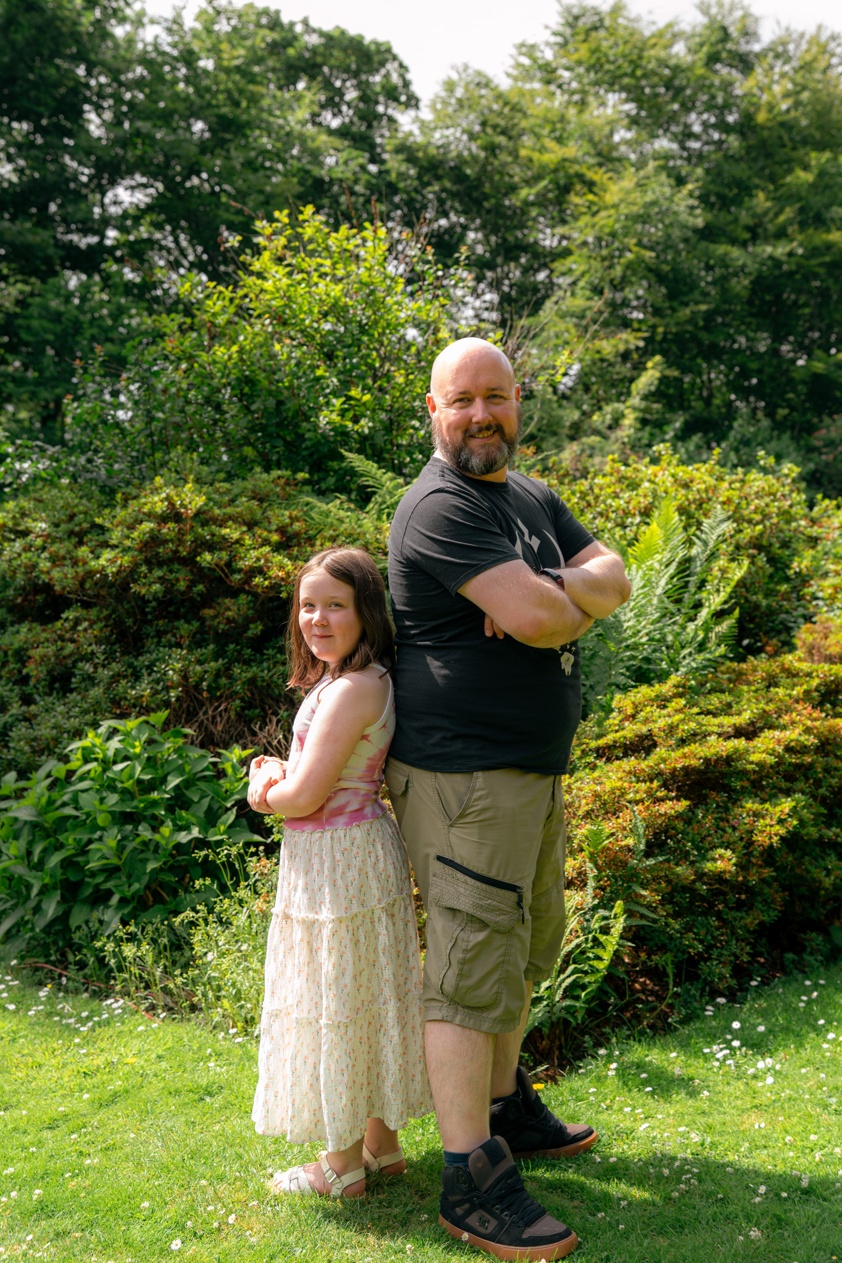 At their Aberdeen family photo shoot, a dad with beard and shaved head stands back to back with his seven year old daughter, seeing who is taller. The background is of sunlit shrubs towering over them both.
