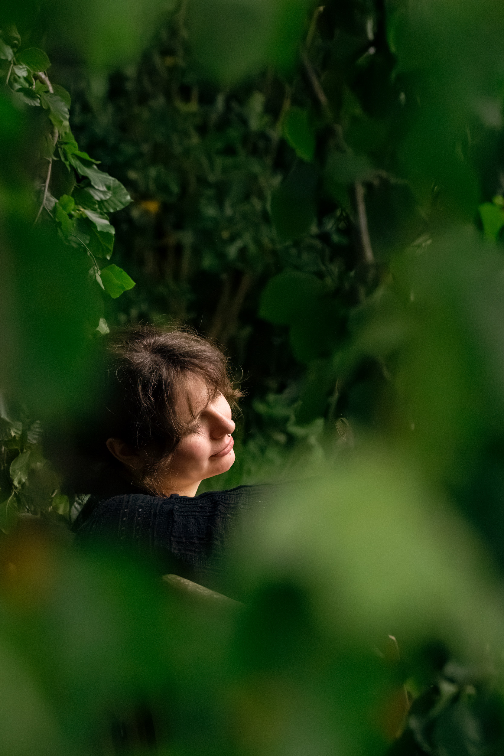 Taken by Aberdeen wedding photographer, Sarah from Windswept Stories, during a couple's photo session, a young woman's softly lit face is shown against a dark backdrop of leaves; her eye lids are lowered and she looks serene. She is framed by soft focus leaves in the foreground.