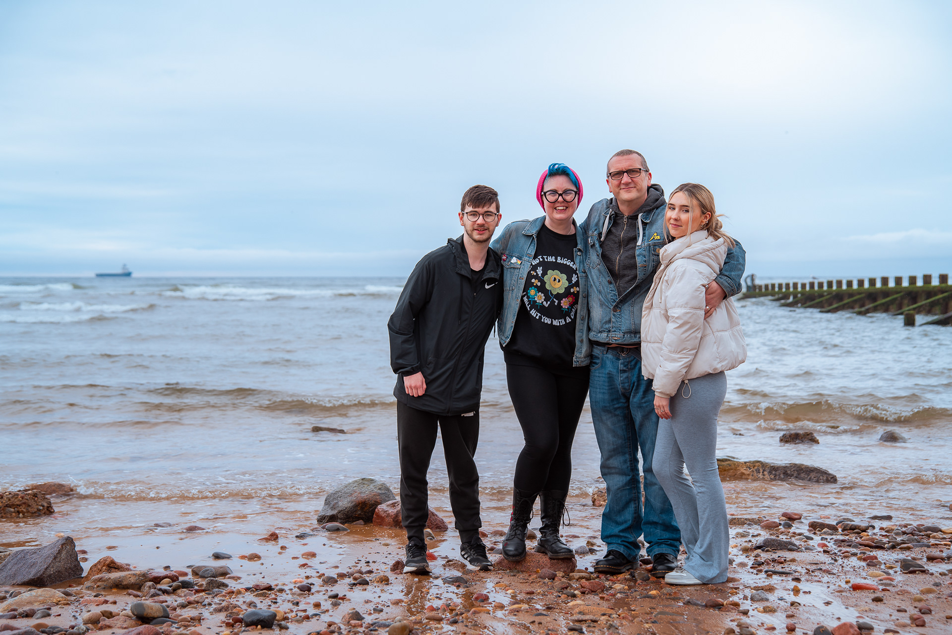 A family of four - two adults, two teenagers - hug and smile directly at the camera on a stony beach, with the North Sea behind them.