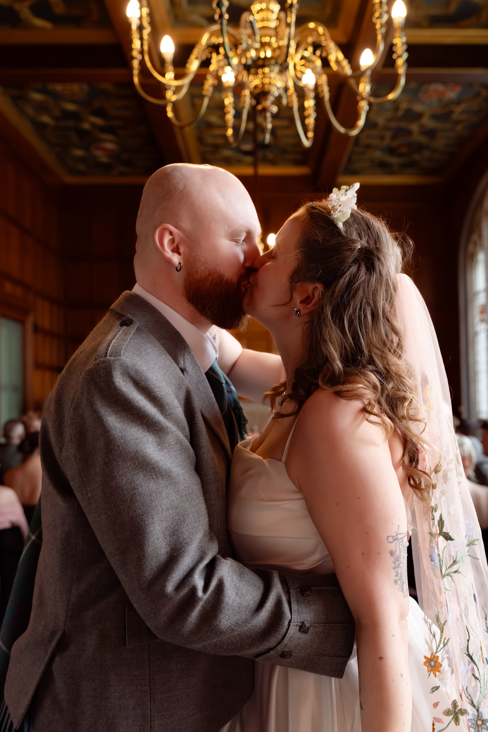 Bride and groom kiss in Marischal College Grant Room, chandelier and ornate ceiling visible behind them.