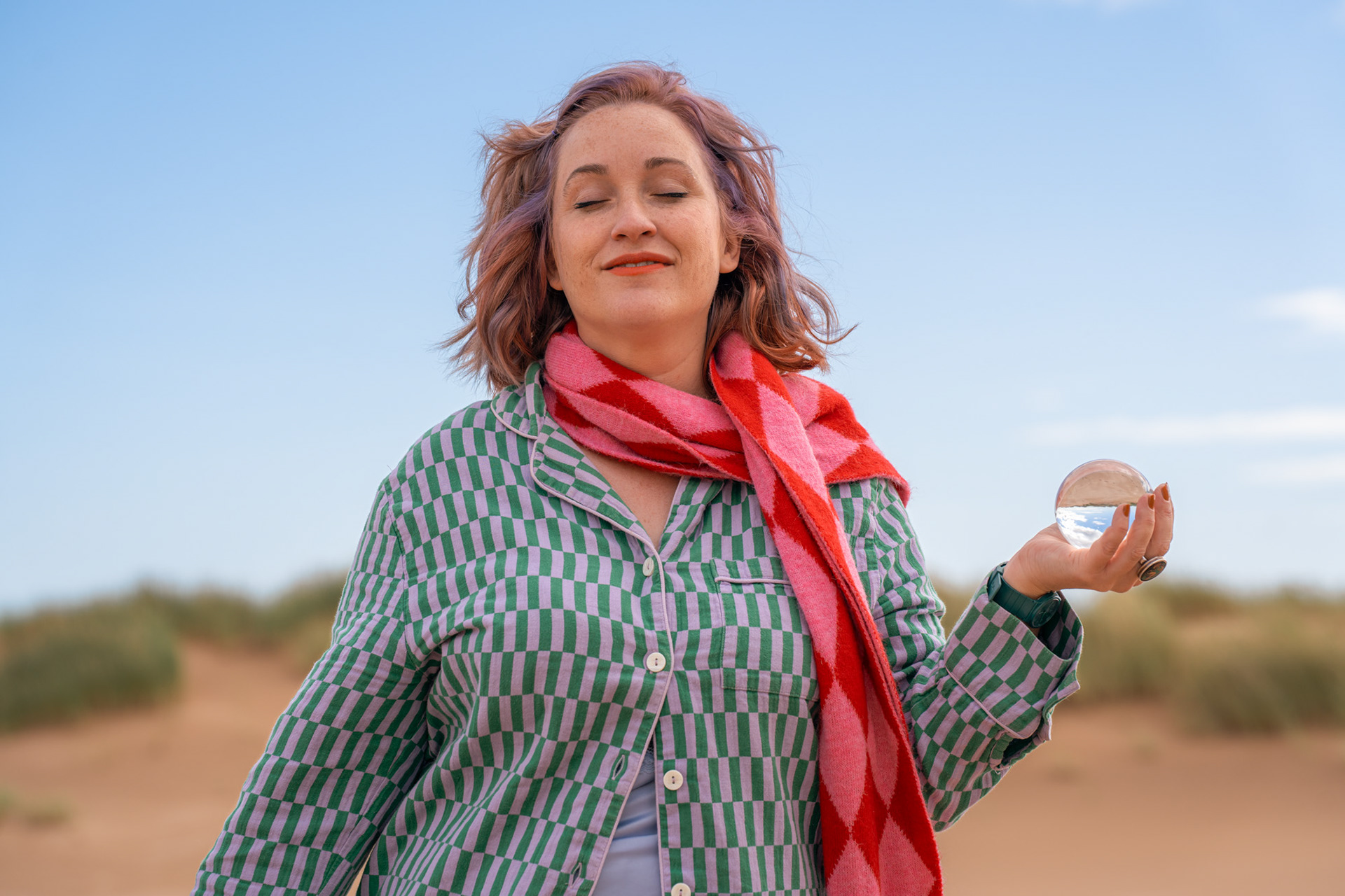 A white woman with ginger and lilac hair and freckles closes her eyes and stands with a crystal ball in one hand, blue skies behind her.