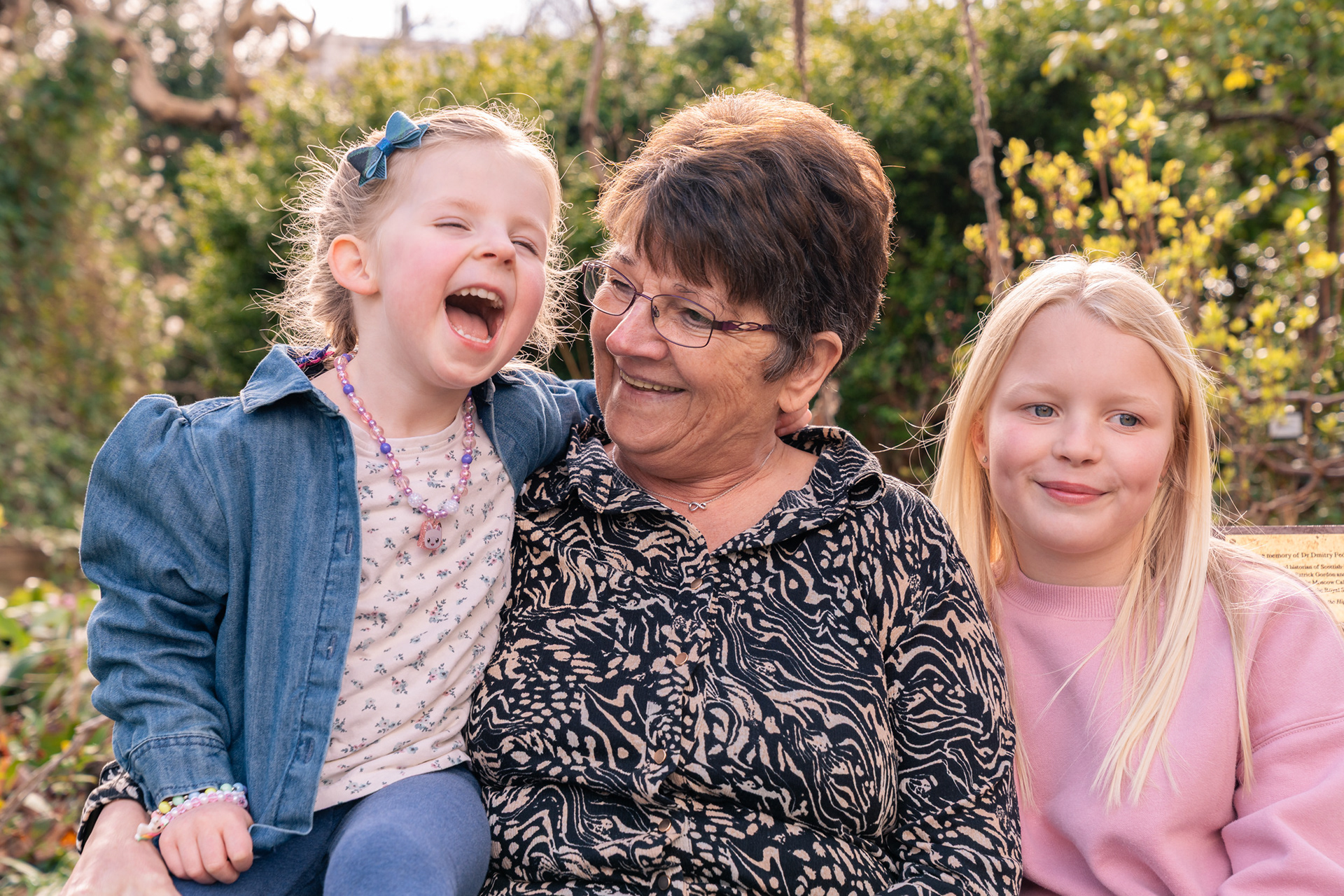 Granny and two granddaughters laugh together, backlit by the sunshine on a spring day, at an outdoor Aberdeen family photo shoot.