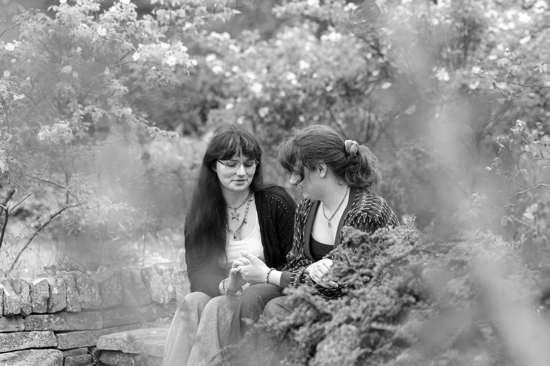 Flowers in the foreground of this black and white image partially obscure the young couple sitting on stone steps in a garden, lightly holding hands and watching their fingers moving as they chat.