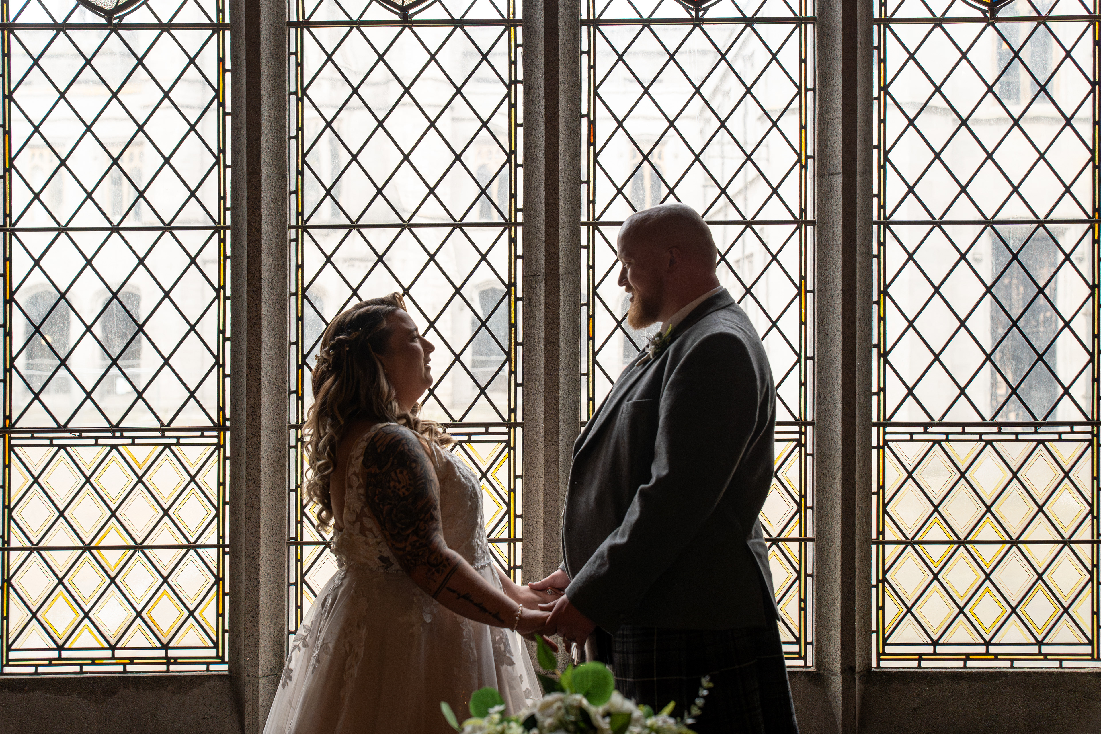 Bride in white and groom in kilt hold hands, silhouetted against stained glass windows in Aberdeen's Marischal College