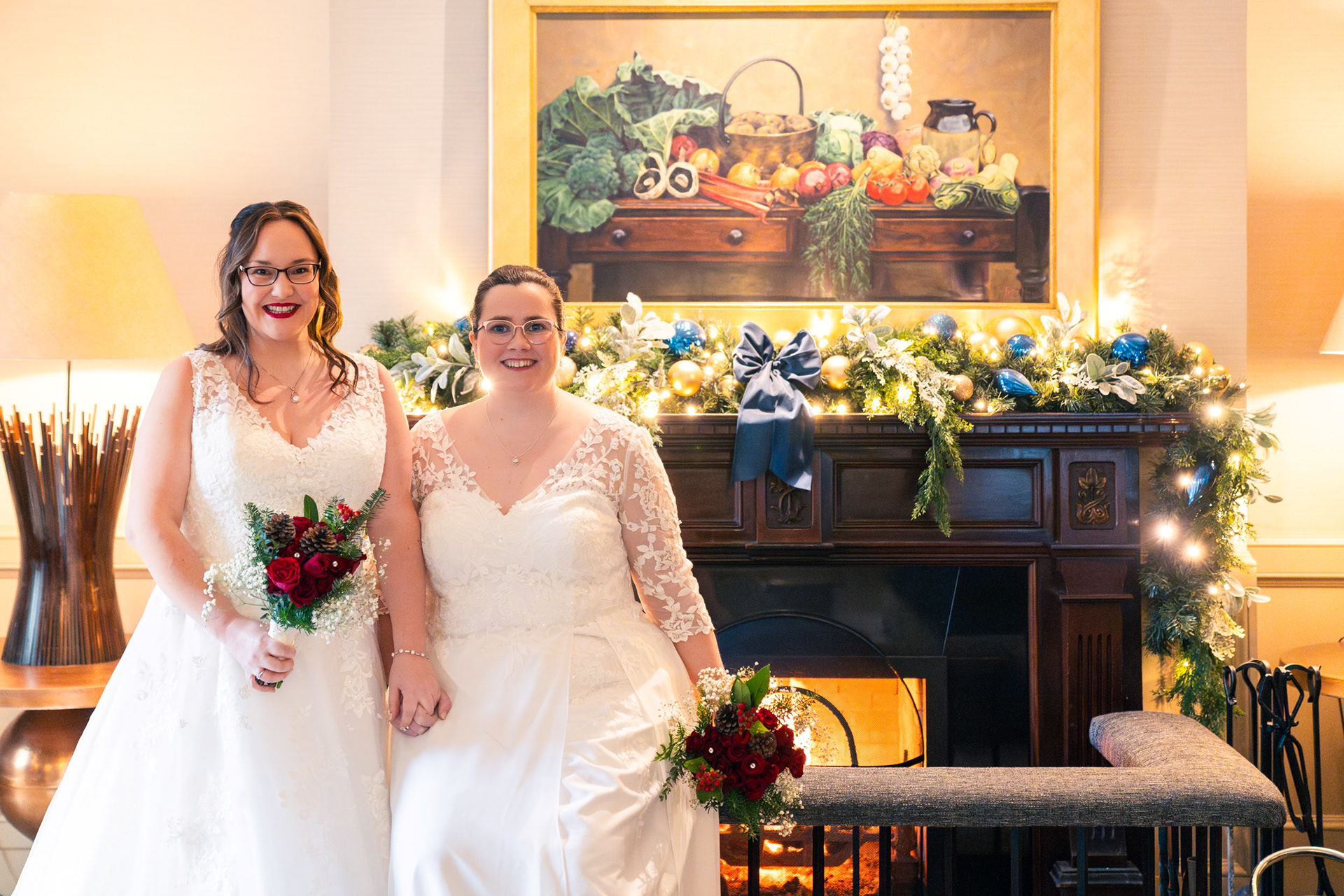 Lesbian Wedding Scotland: Two brides in white dresses, both brunette and wearing glasses, hold bouquets of red roses and sit in front of a roaring fire surrounded by Christmas lights and baubles in Aberdeen's Marcliffe Hotel.