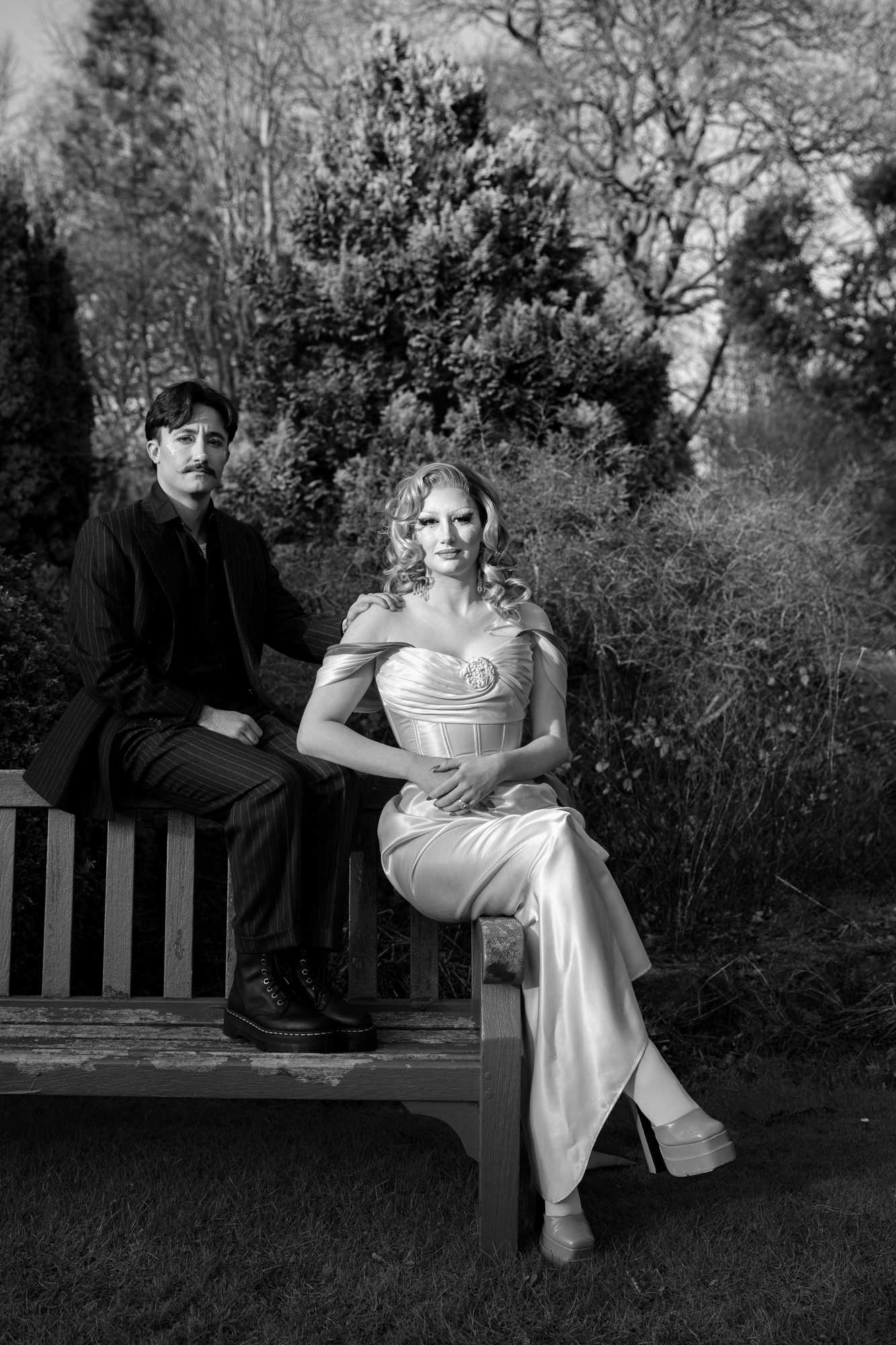A couple in drag pose together during their Aberdeen couple's photo session. They/them is wearing a pinstripe suit and has short, dark hair and a moustache. She/her is in a glamorous pink and green dress and high green heels with dramatic makeup.  She sits on the arm of a bench; he sits on the back of the bench, one hand on her shoulder.