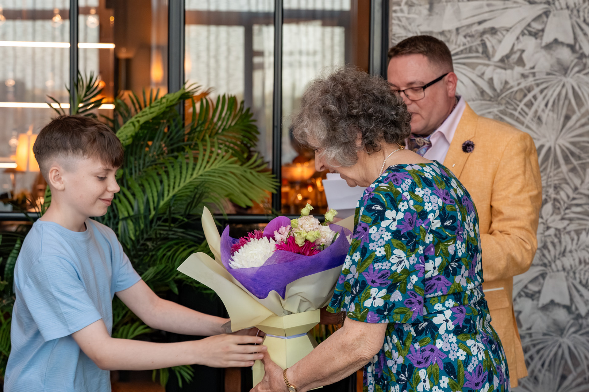 Aberdeen Photographer: A boy in a blue T-shirt hands a colourful bouquet of flowers to an older woman in a blue and purple floral dress.