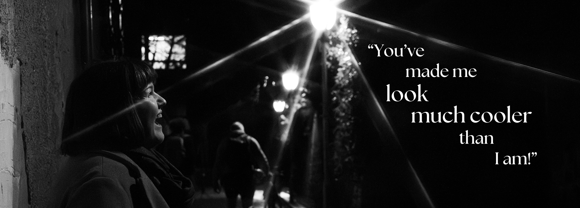 Black and white image of laughing woman with dark bob. Rays of light from a nearby streetlight bisect the image. In a dark section, the caption reads "You've made me look much cooler than I am!"